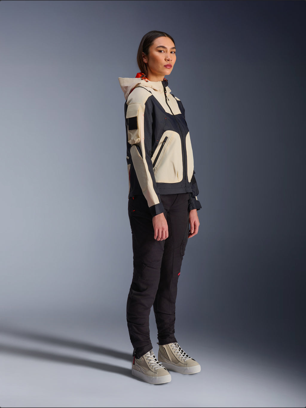 Kurtka Alpinestars Molly Air Jacket w kolorze Black Almond, noszona przez motocyklistkę z czarnymi spodniami i kremowymi wysokimi sneakersami, charakteryzująca się dwukolorowym designem z czarnymi i beżowymi panelami, zintegrowanym kapturem i nowoczesnym stylem miejskim.