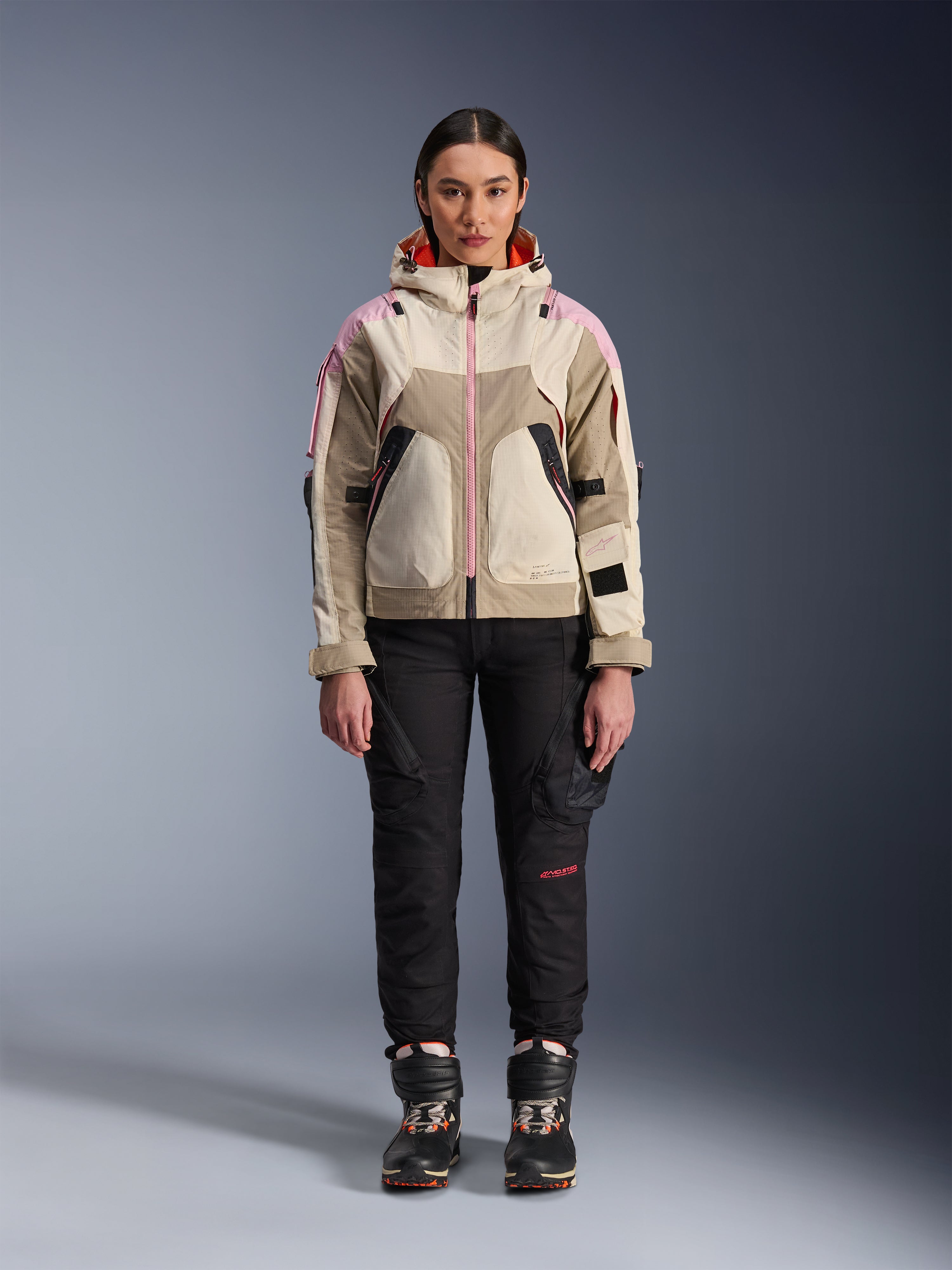 Modelka w kurtce Alpinestars Molly Air Jacket w kolorze Sand Pink Almond, czarne techniczne spodnie motocyklowe i buty, widok z przodu na całą sylwetkę, miejska kurtka motocyklowa o designie inspirowanym stylem utility i różowymi akcentami