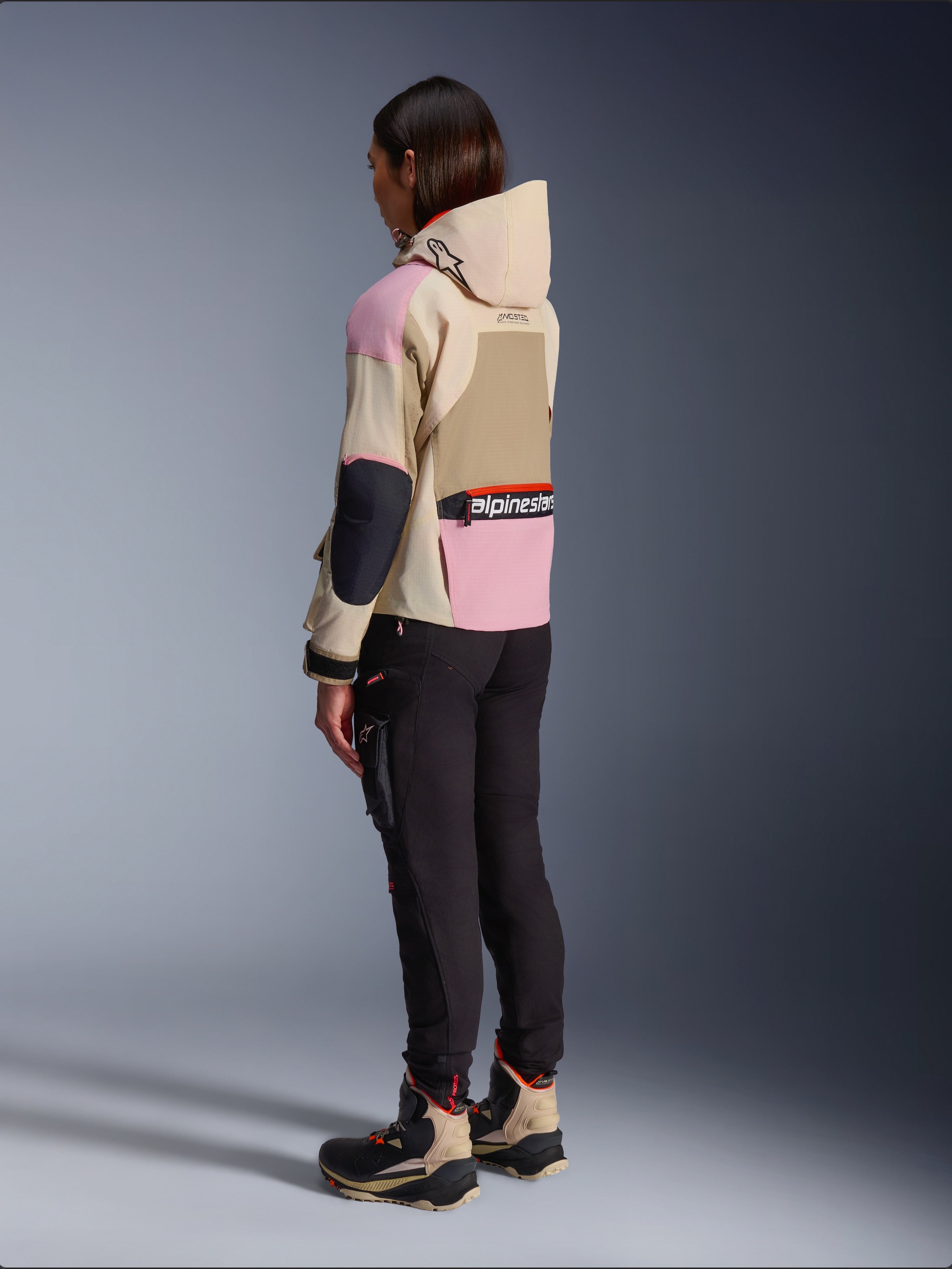 Motocyklistka w kurtce Alpinestars Molly Air Jacket w kolorze Sand Pink Almond, wielokolorowy beżowo-różowy design, widok z boku i z tyłu, czarne spodnie motocyklowe i dopasowane buty techniczne, miejski styl motocyklowy z kapturem