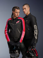 Alpinestars Stella T-Dyno Air Jacket na kobiecie w kolorze czarnym, jaskrawoczerwonym i fuksji, w zestawieniu z męską czarną kurtką Dyno Air Jacket, oboje motocykliści trzymają kaski motocyklowe, ujęcie studyjne współczesnej miejskiej odzieży motocyklowej