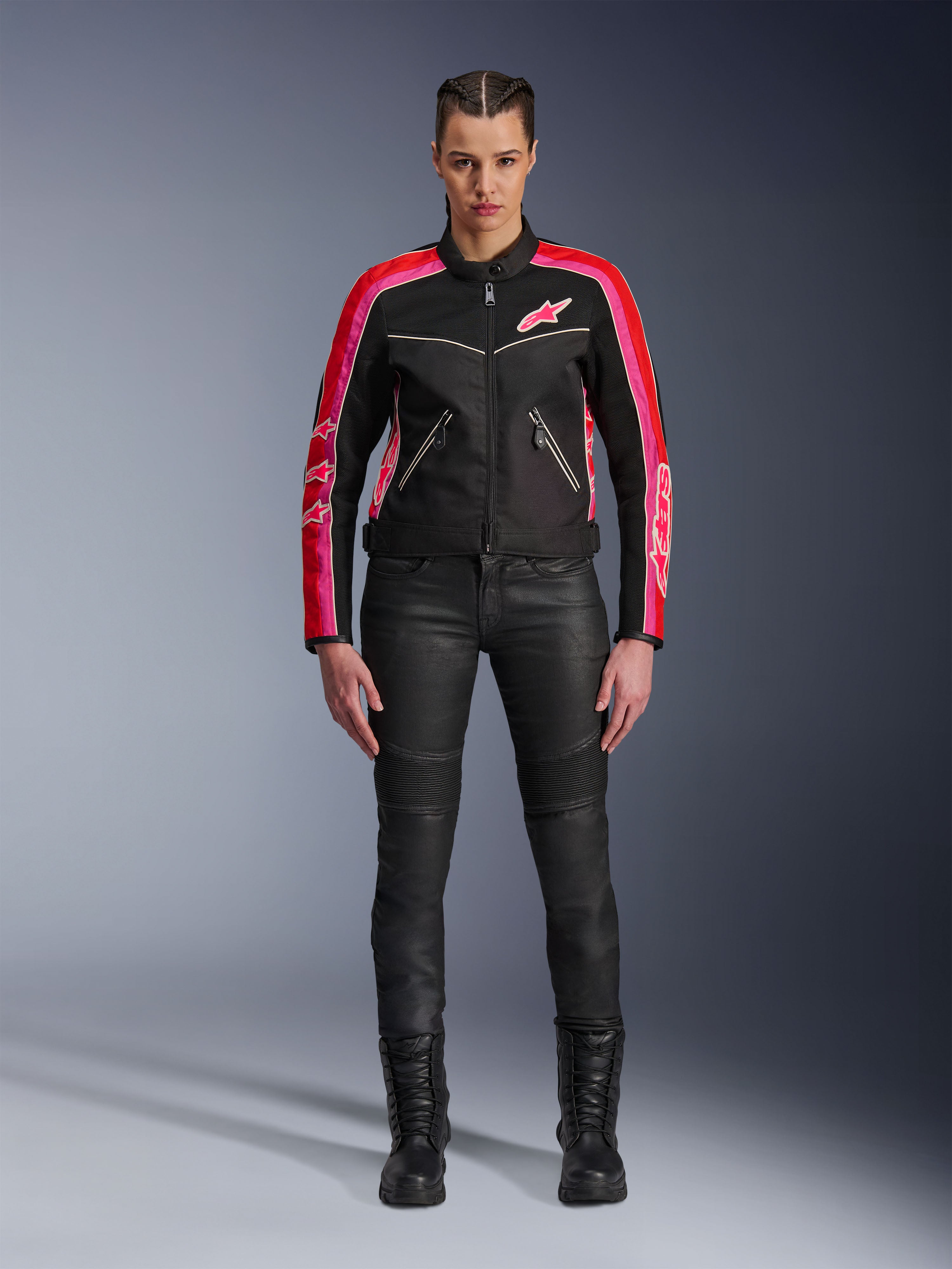 Motocyklistka w kurtce Alpinestars Stella T-Dyno Air, czarna z jaskrawoczerwonymi i fuksjowymi akcentami, zestawiona z czarnymi spodniami motocyklowymi i sznurowanymi butami, panele z siatki 3D, dopasowany krój, różowe logo gwiazdy, miejski strój motocyklowy