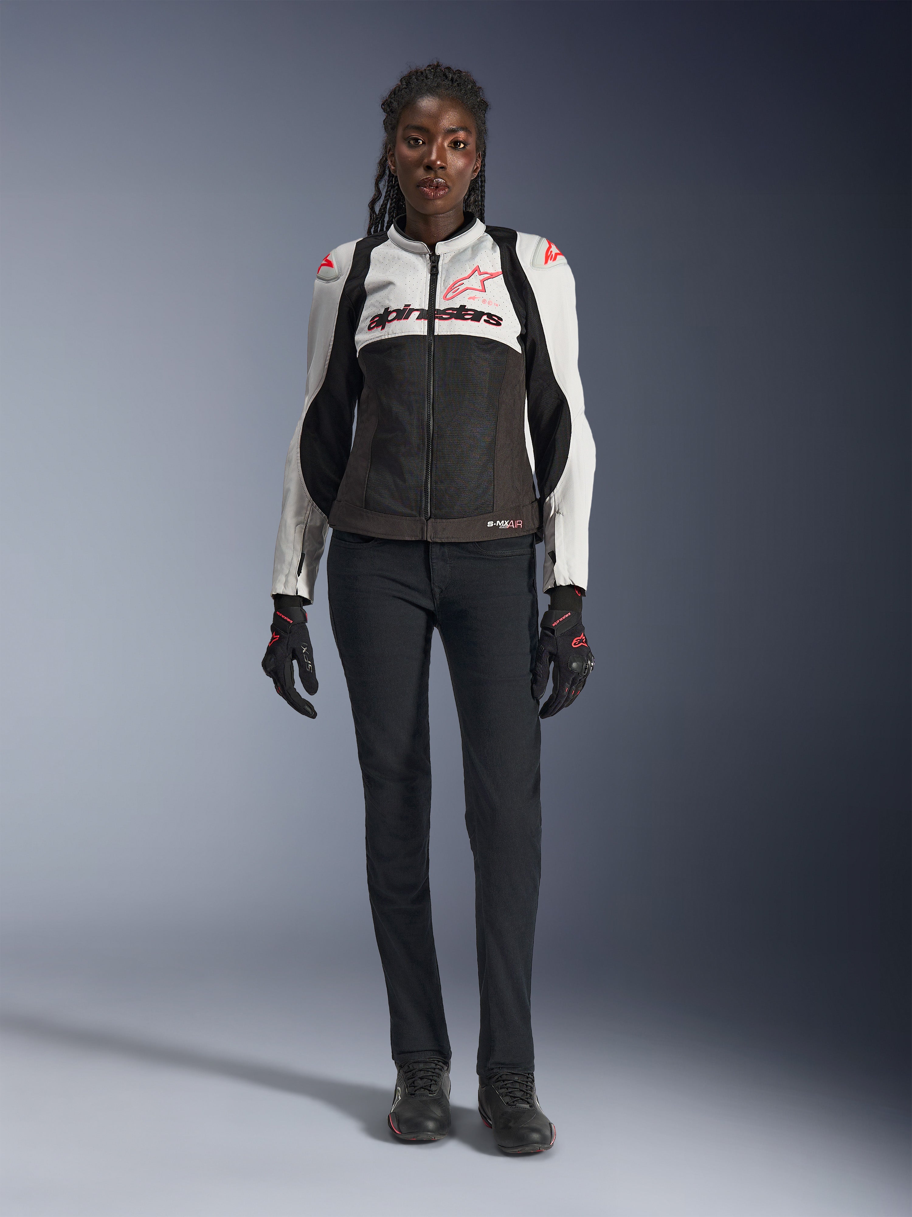 Kurtka Alpinestars Stella SMX Air, Ice Gray Black i Coral Fluo, prezentowana na modelce w czarnych spodniach i rękawicach motocyklowych, z siatkową konstrukcją i ochronnymi sliderami na ramionach, sportowa odzież motocyklowa dla kobiet