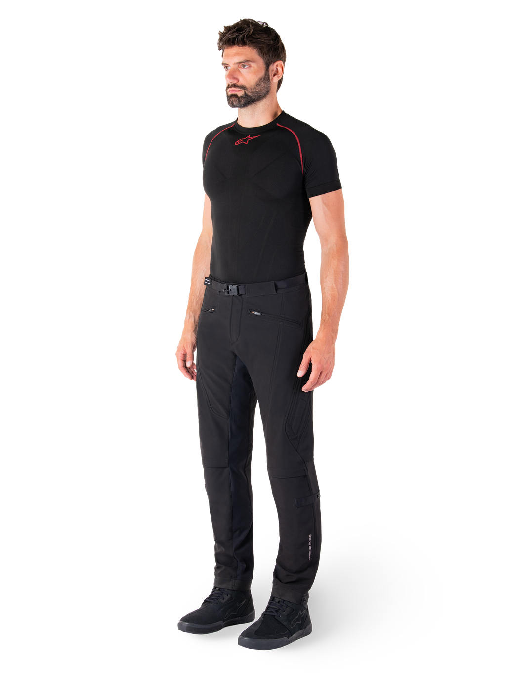 Alpinestars Alden Pants, miejskie spodnie motocyklowe, czarne, tekstylny softshell o regularnym kroju, prezentowane na modelu z czarną warstwą bazową i butami do jazdy, wyposażone w pasek w talii z zapięciem magnetycznym i kieszenie na zamek.
