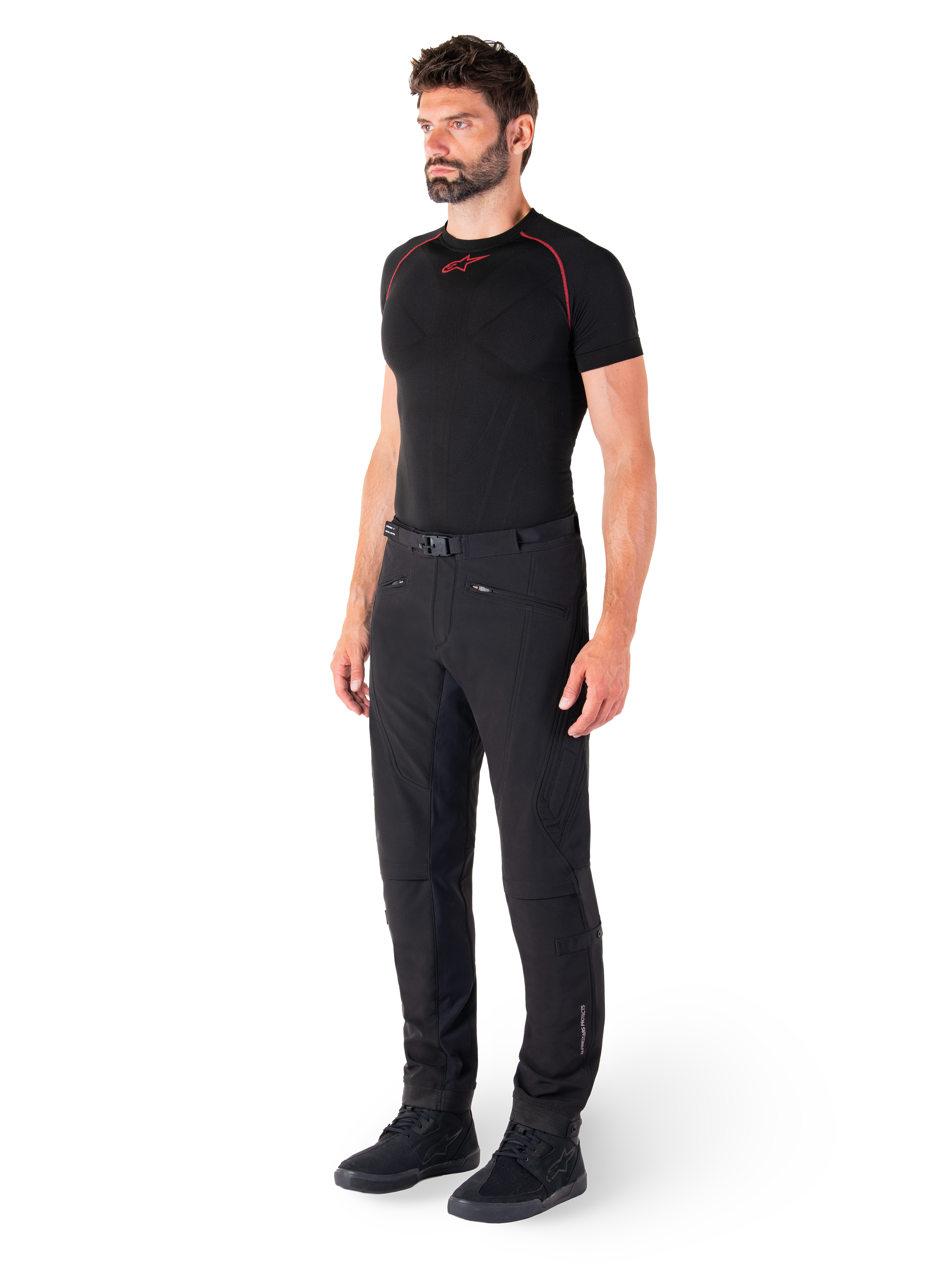 Alpinestars Alden Pants, miejskie spodnie motocyklowe, czarne, tekstylny softshell o regularnym kroju, prezentowane na modelu z czarną warstwą bazową i butami do jazdy, wyposażone w pasek w talii z zapięciem magnetycznym i kieszenie na zamek.