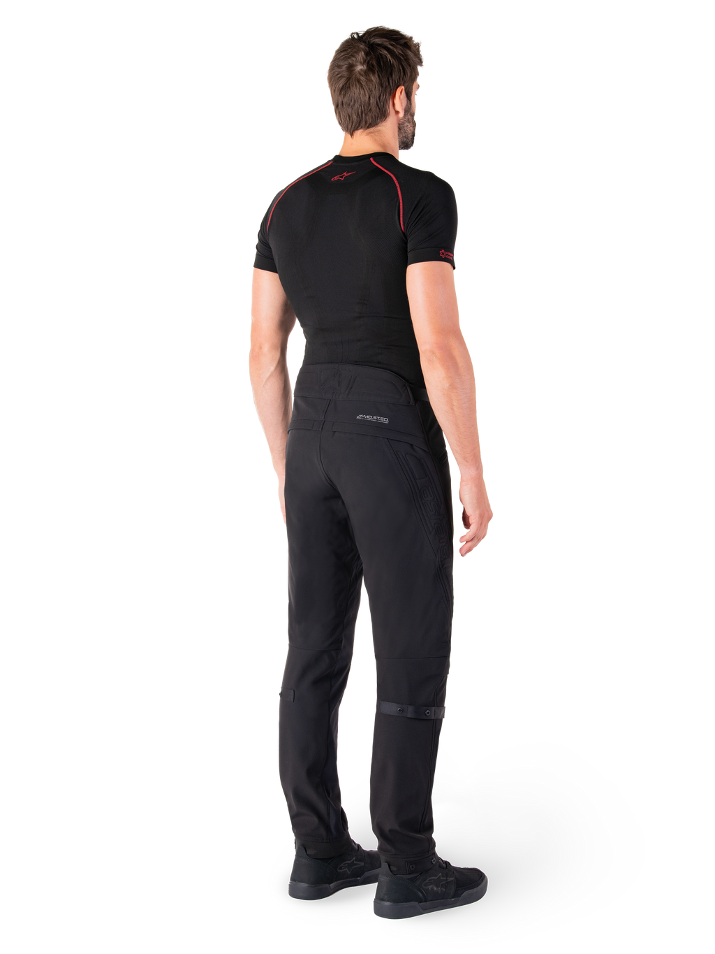 Alpinestars Alden Pants, czarne, miejskie spodnie motocyklowe widoczne od tyłu na modelu, noszone z czarną koszulką z krótkim rękawem i czarnymi butami do jazdy, z pikowanymi detalami i odblaskowym logo MO.ST.EQ. na tylnym karczku.