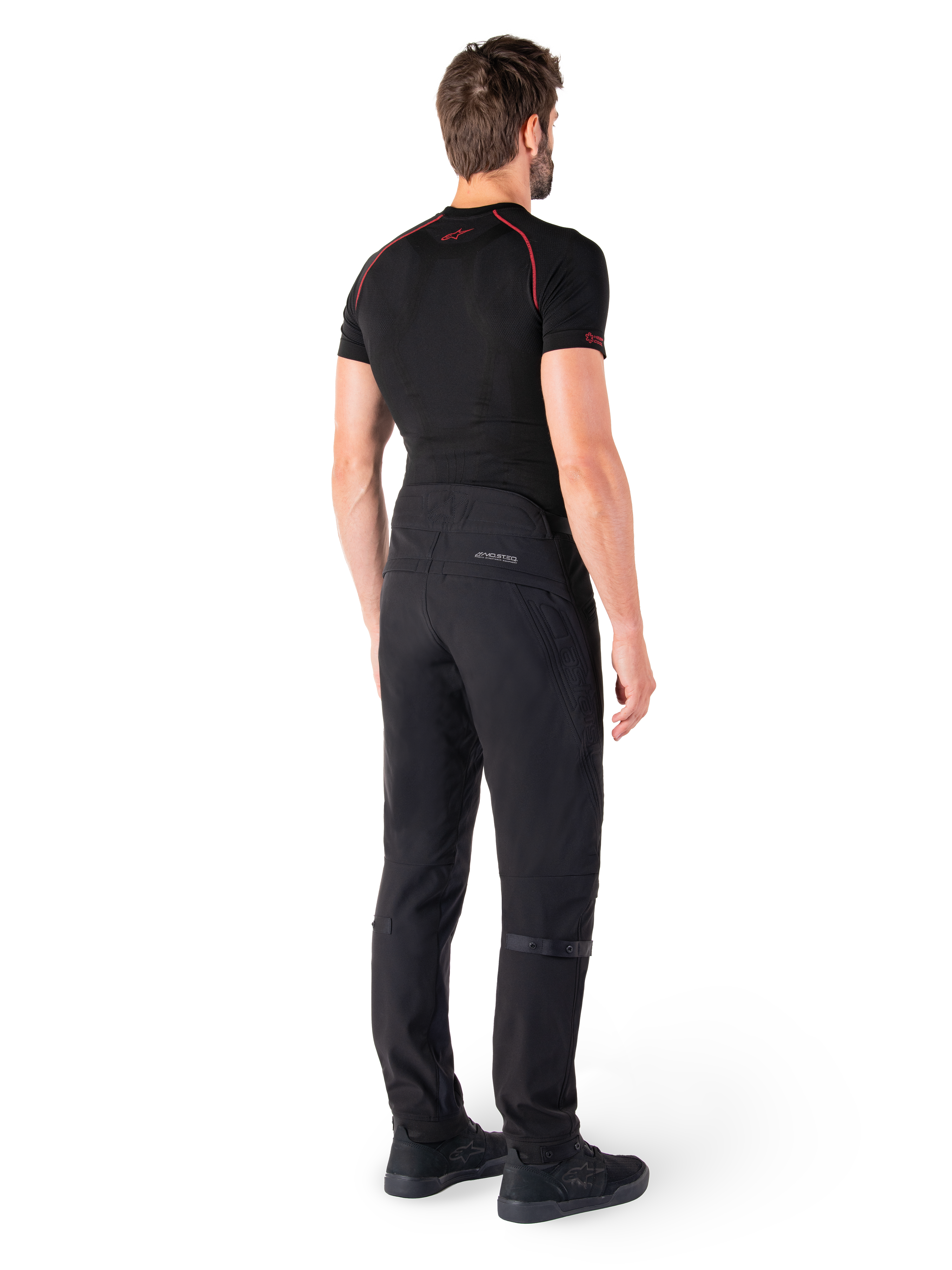 Alpinestars Alden Pants, czarne, miejskie spodnie motocyklowe widoczne od tyłu na modelu, noszone z czarną koszulką z krótkim rękawem i czarnymi butami do jazdy, z pikowanymi detalami i odblaskowym logo MO.ST.EQ. na tylnym karczku.