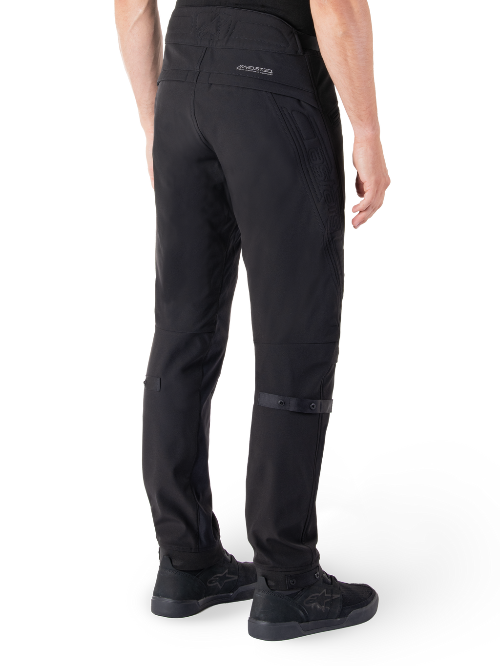 Spodnie Alpinestars Alden, miejskie spodnie motocyklowe, czarne, widok z tyłu ukazujący techniczny materiał softshell z pikowanymi detalami, odblaskowe logo MO.ST.EQ. na tylnym karczku oraz regulowane paski na łydkach, zestawione z czarnymi butami motocyklowymi