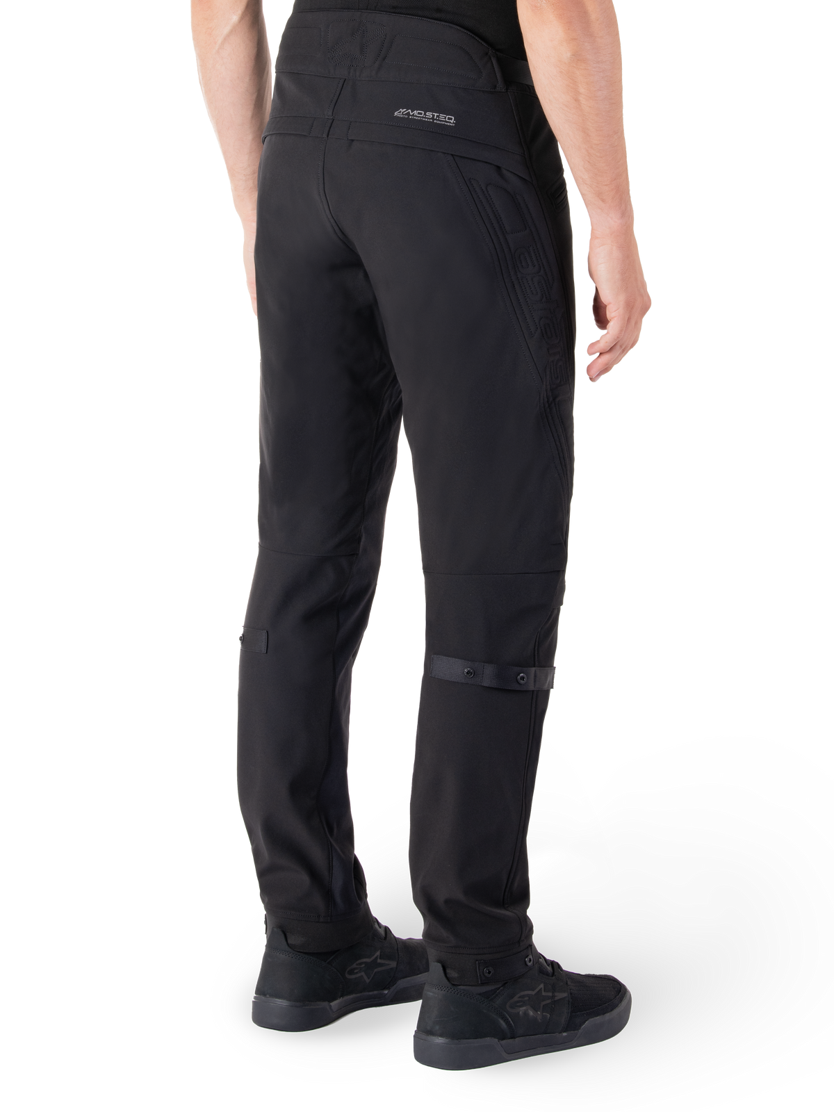 Spodnie Alpinestars Alden, miejskie spodnie motocyklowe, czarne, widok z tyłu ukazujący techniczny materiał softshell z pikowanymi detalami, odblaskowe logo MO.ST.EQ. na tylnym karczku oraz regulowane paski na łydkach, zestawione z czarnymi butami motocyklowymi