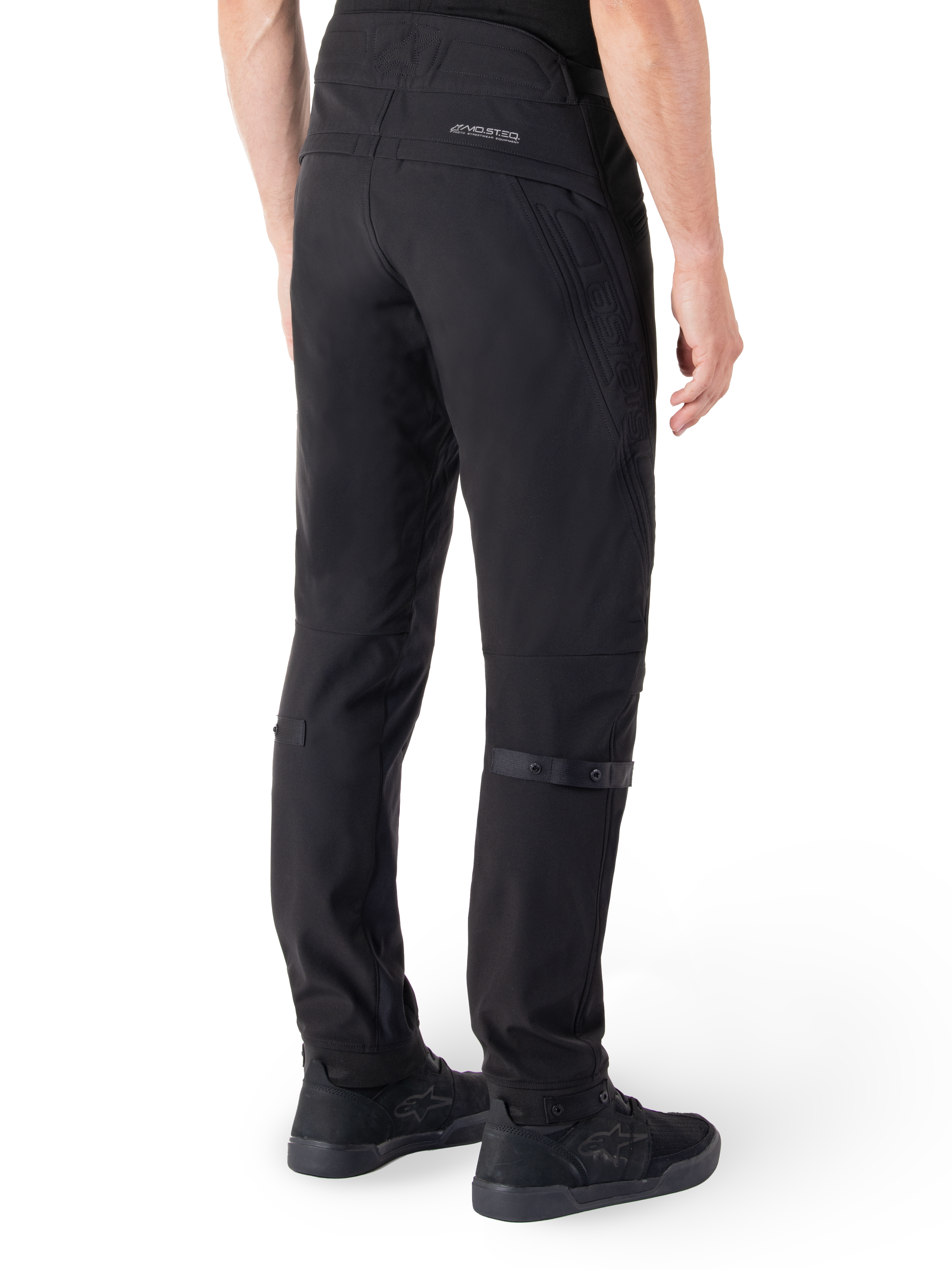 Spodnie Alpinestars Alden, miejskie spodnie motocyklowe, czarne, widok z tyłu ukazujący techniczny materiał softshell z pikowanymi detalami, odblaskowe logo MO.ST.EQ. na tylnym karczku oraz regulowane paski na łydkach, zestawione z czarnymi butami motocyklowymi