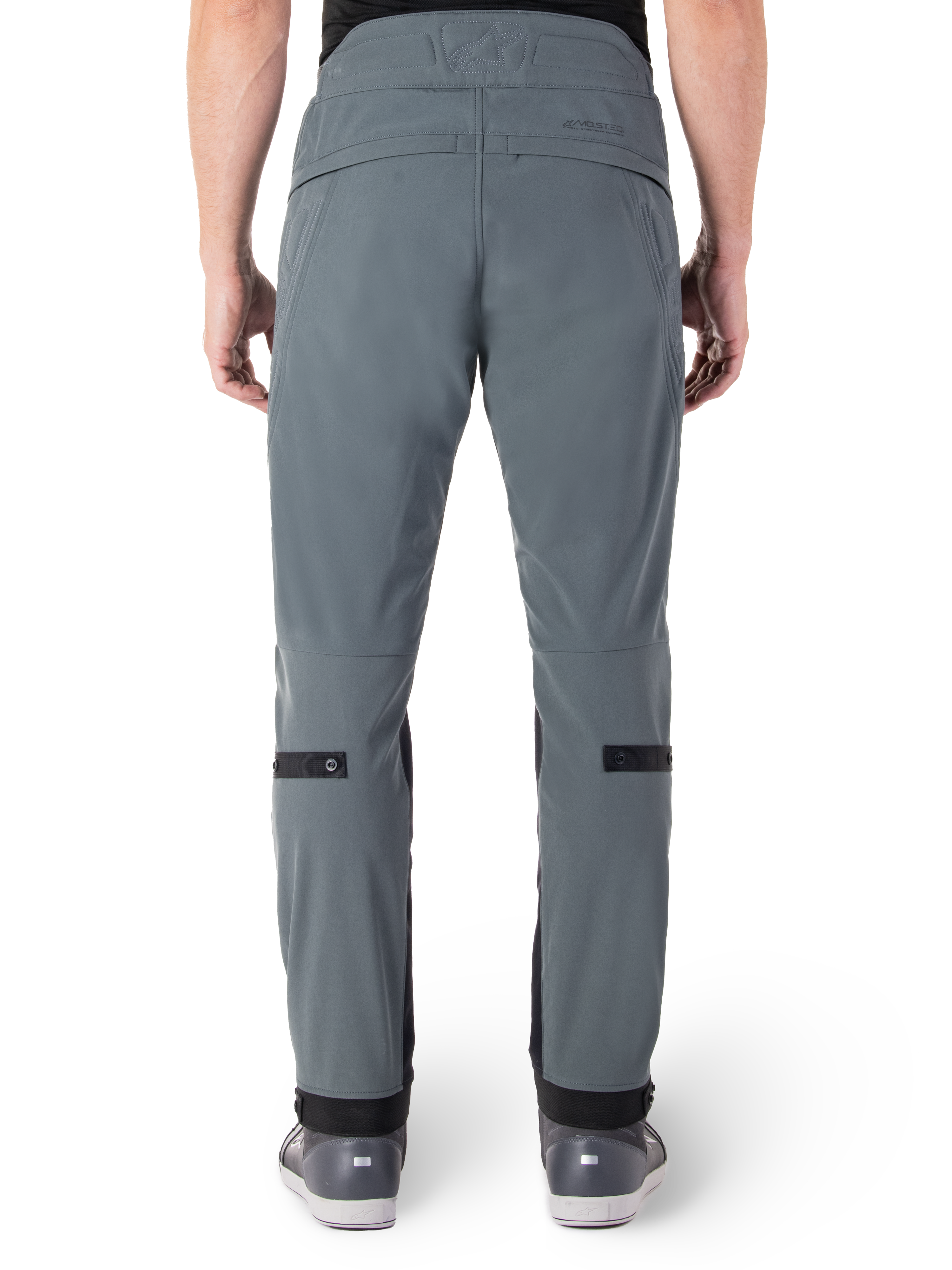 Alpinestars Alden Pants, Urban Gray, widok z tyłu męskich miejskich spodni motocyklowych, wąski krój poniżej kolana z czarnymi paskami do regulacji na łydkach, wysoki stan z napisem MO.ST.EQ. na tylnym karczku, noszone z szarymi butami do jazdy