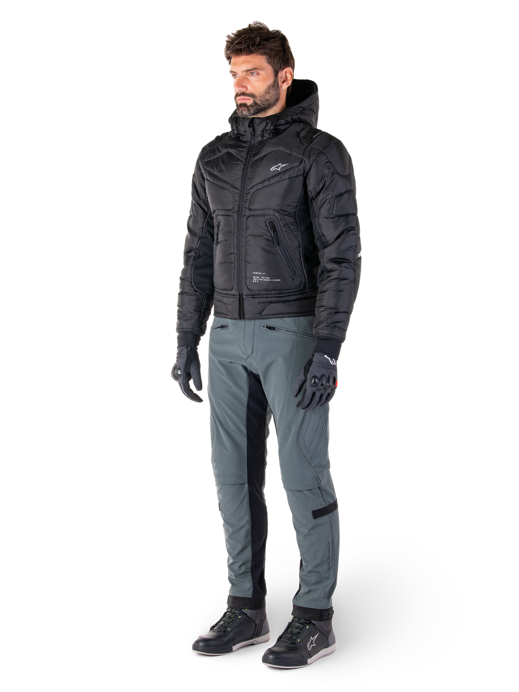 Spodnie Alpinestars Alden w kolorze Urban Gray, prezentowane przez modela w czarnej pikowanej kurtce z kapturem, rękawicach i butach motocyklowych, pikowane detale po bokach i czarne panele po wewnętrznej stronie nogawek, miejska odzież motocyklowa inspirowana stylem streetwear