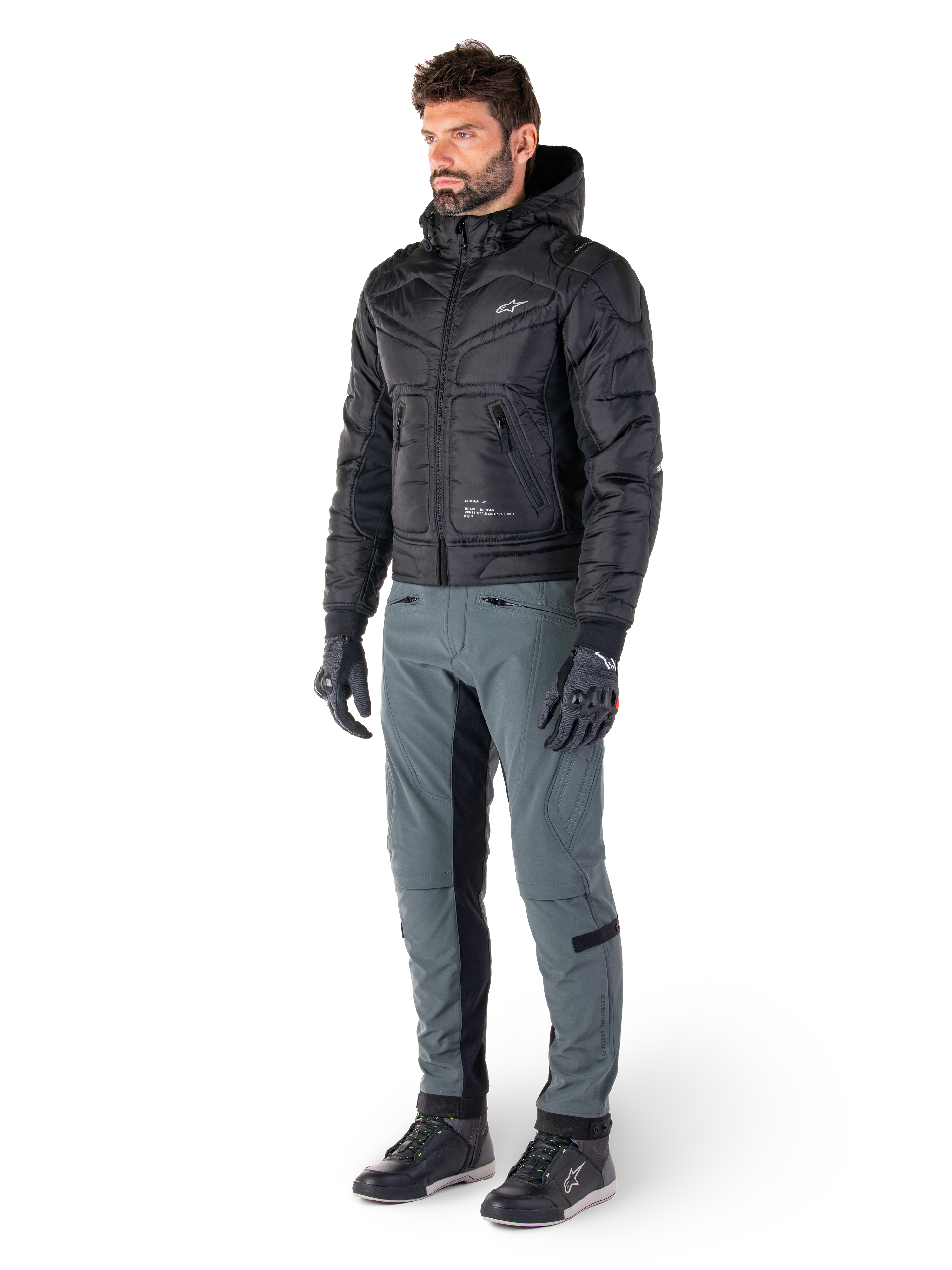 Spodnie Alpinestars Alden w kolorze Urban Gray, prezentowane przez modela w czarnej pikowanej kurtce z kapturem, rękawicach i butach motocyklowych, pikowane detale po bokach i czarne panele po wewnętrznej stronie nogawek, miejska odzież motocyklowa inspirowana stylem streetwear