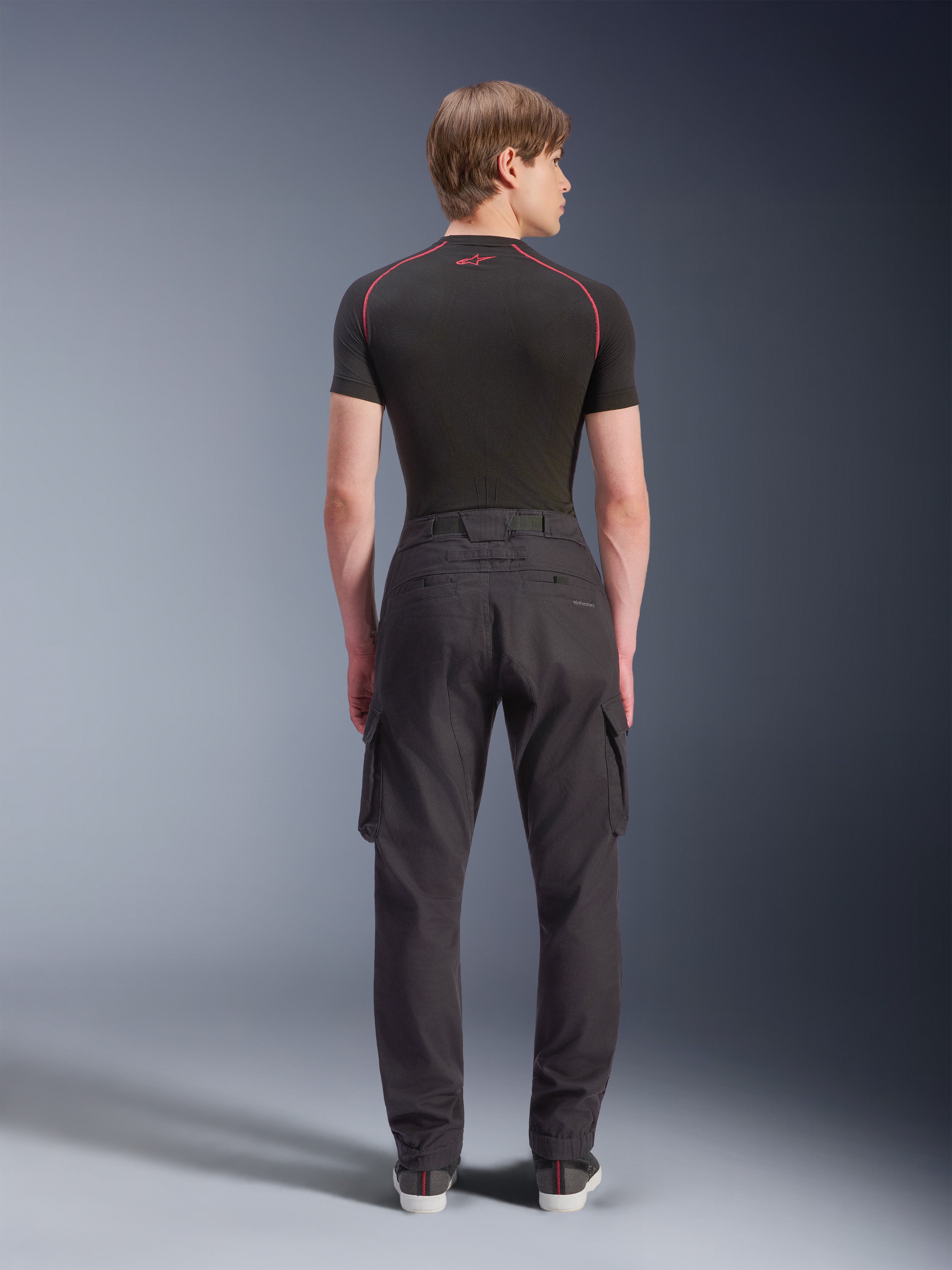 Alpinestars Flex-AST Cargo Pants - Short, czarne, miejskie spodnie motocyklowe widoczne od tyłu, z bocznymi kieszeniami cargo i elastycznymi mankietami, noszone z czarną warstwą bazową z krótkim rękawem i czerwonymi kontrastowymi szwami.
