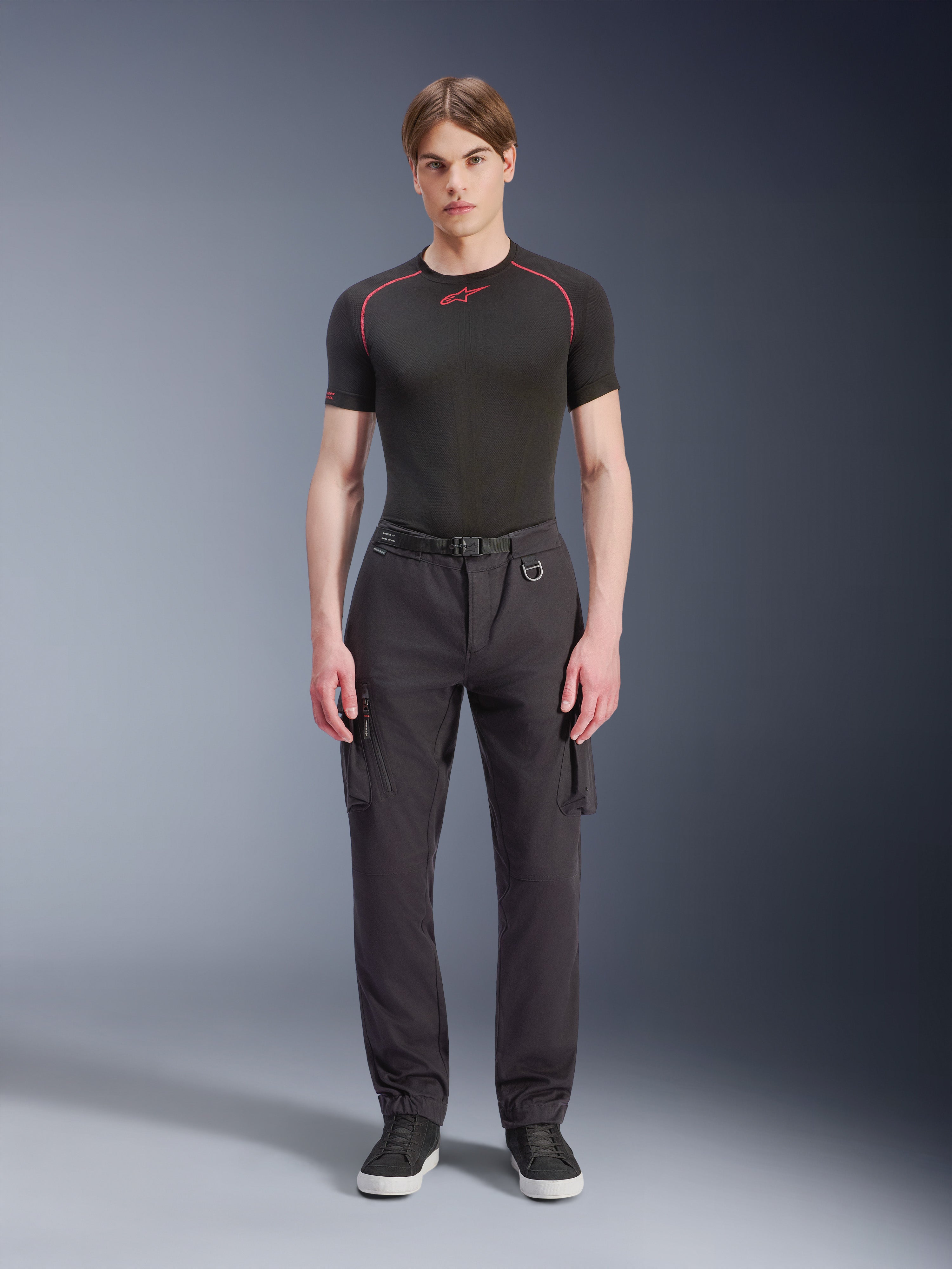 Alpinestars Flex-AST Cargo Pants, czarne, miejskie spodnie motocyklowe prezentowane przez modela w czarnej koszulce z krótkim rękawem i sneakersach, charakteryzujące się standardowym zwężanym krojem, kieszeniami cargo i zintegrowaną klamrą paska