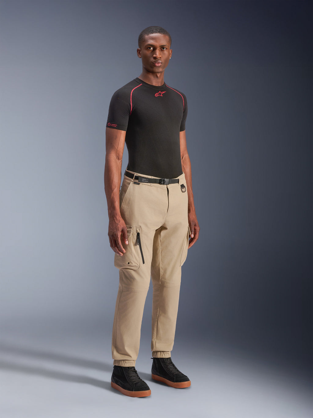 Alpinestars Flex-AST Cargo Pants - Short, jasnobeżowe miejskie spodnie motocyklowe, noszone przez modela z czarną warstwą bazową i sneakersami, o regularnym zwężanym kroju, z elastycznymi mankietami i wielokieszeniowym designem cargo