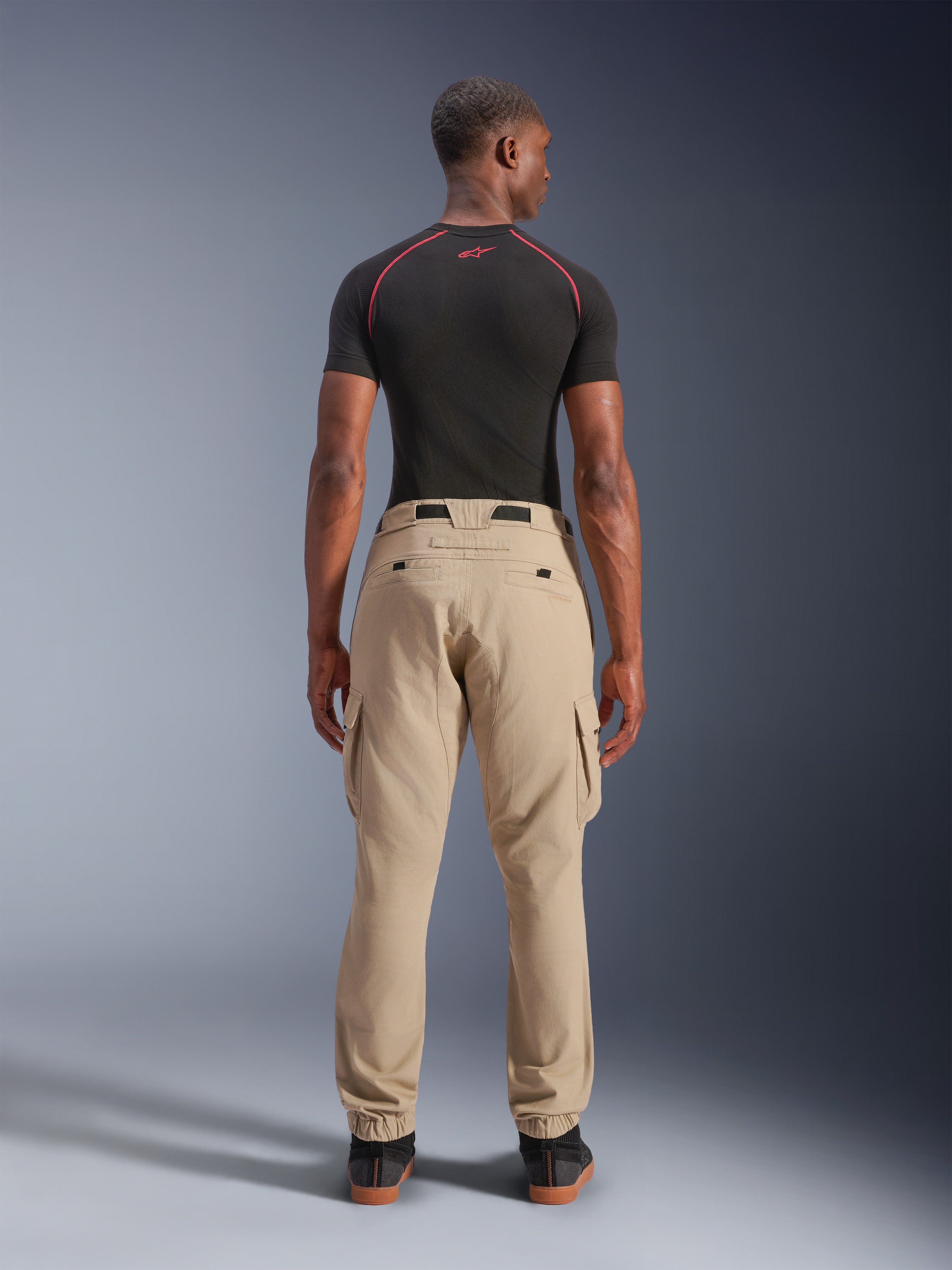 Alpinestars Flex-AST Cargo Pants, jasnobeżowe, widok z tyłu na modelu, regularny zwężany krój z bocznymi kieszeniami cargo i elastycznymi mankietami, zestawione z czarną warstwą bazową z krótkim rękawem z czerwonymi akcentami, miejska odzież motocyklowa