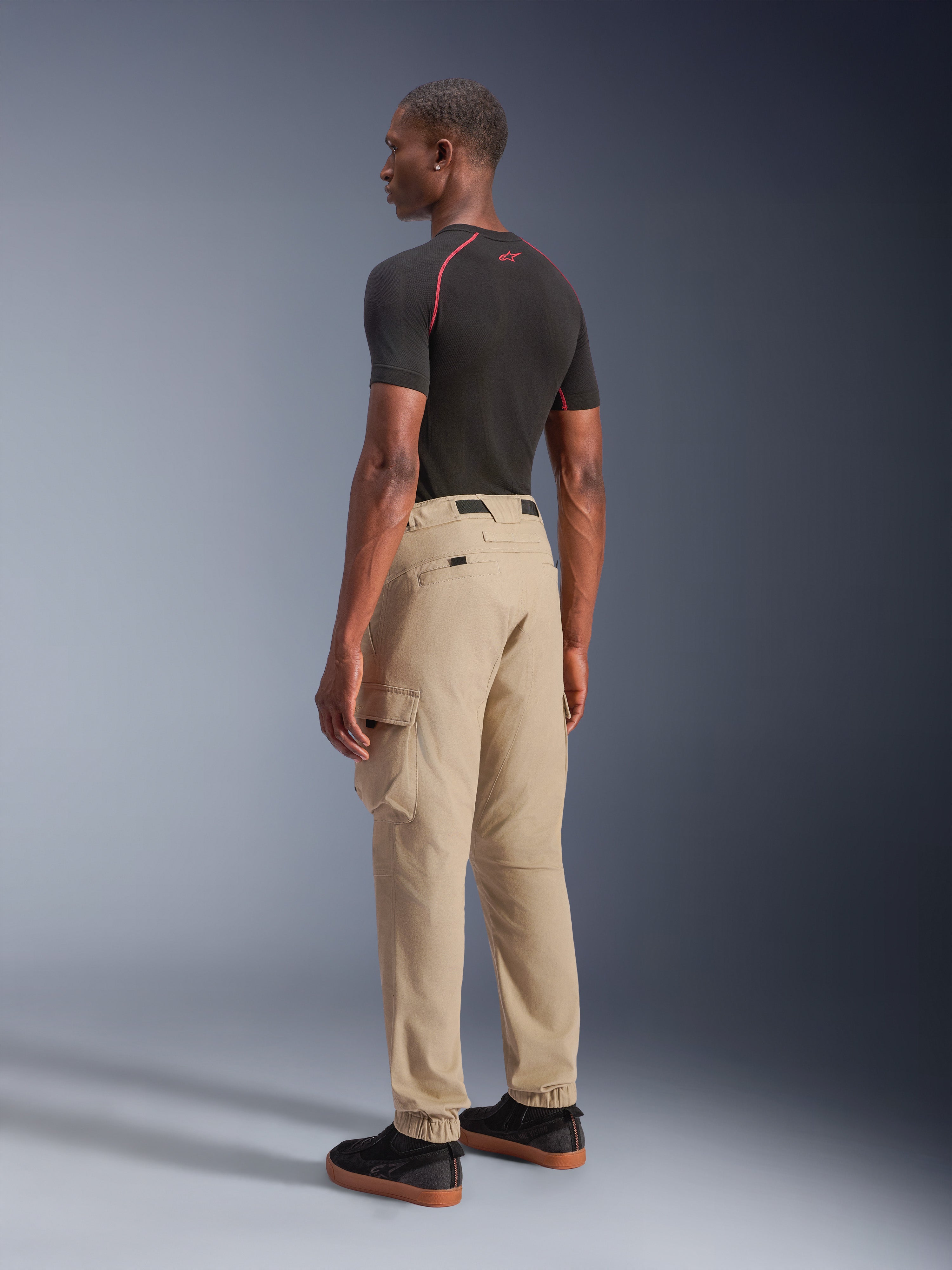 Alpinestars Flex-AST Cargo Pants - Short, miejskie spodnie motocyklowe w kolorze jasnobeżowym, widok z tyłu z boku pokazujący boczne kieszenie cargo i elastyczne mankiety, stylizowane z czarną koszulką z krótkim rękawem i czarnymi butami casualowymi