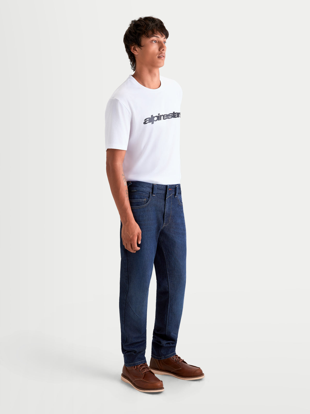 Alpinestars Flex Ast 7 Denim spodnie motocyklowe męskie miejskie, ciemnoniebieskie, widok z boku na modela w białym t-shircie i brązowych butach, jednowarstwowe jeansy motocyklowe o elastycznym komforcie