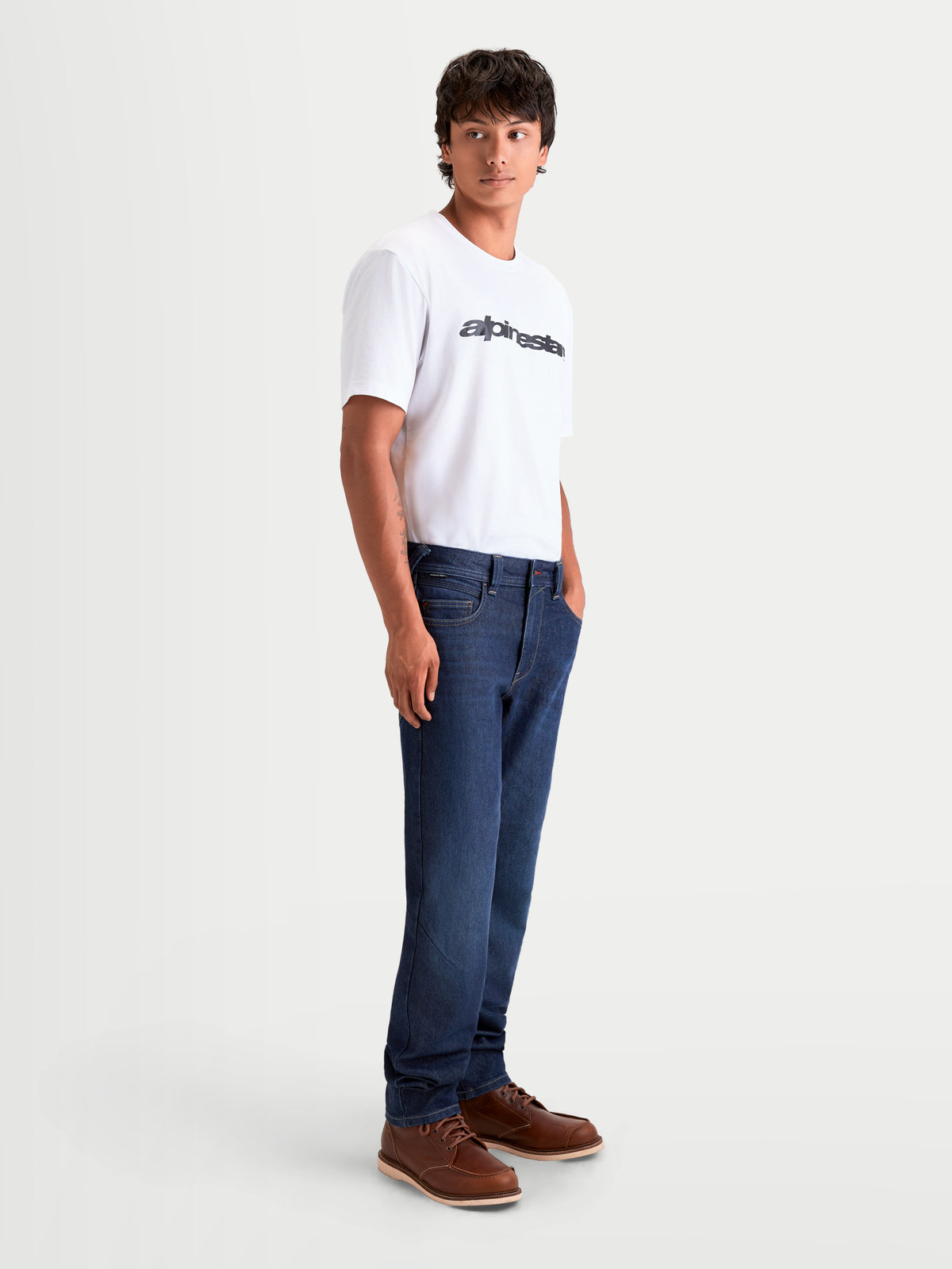 Alpinestars Flex Ast 7 Denim, męskie jeansy motocyklowe urban contemporary w kolorze ciemnoniebieskim, widok z boku na modela w białym T-shircie i brązowych butach, konstrukcja z pojedynczą warstwą CORDURA® z certyfikatem AA