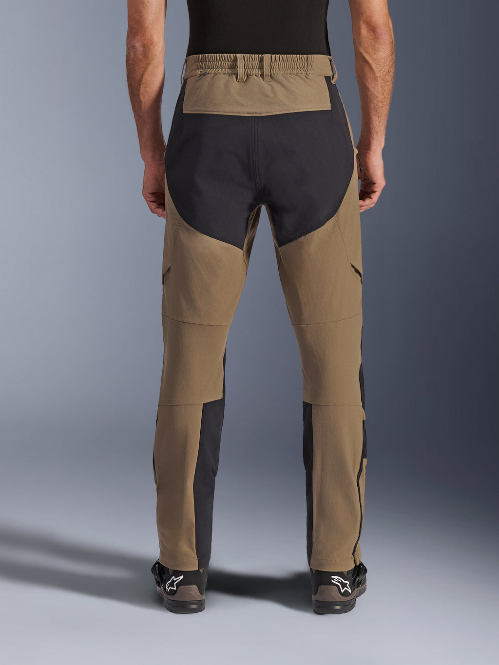 Alpinestars Flex-AST Explorer Pants, motocyklowe spodnie adventure, Walnut Black, widok z tyłu na motocykliście, czarne panele wzmacniające ripstop na siedzeniu i wewnętrznej stronie nogawek, elastyczny pas, długie zamki na dole nogawek, pasujące buty adventure