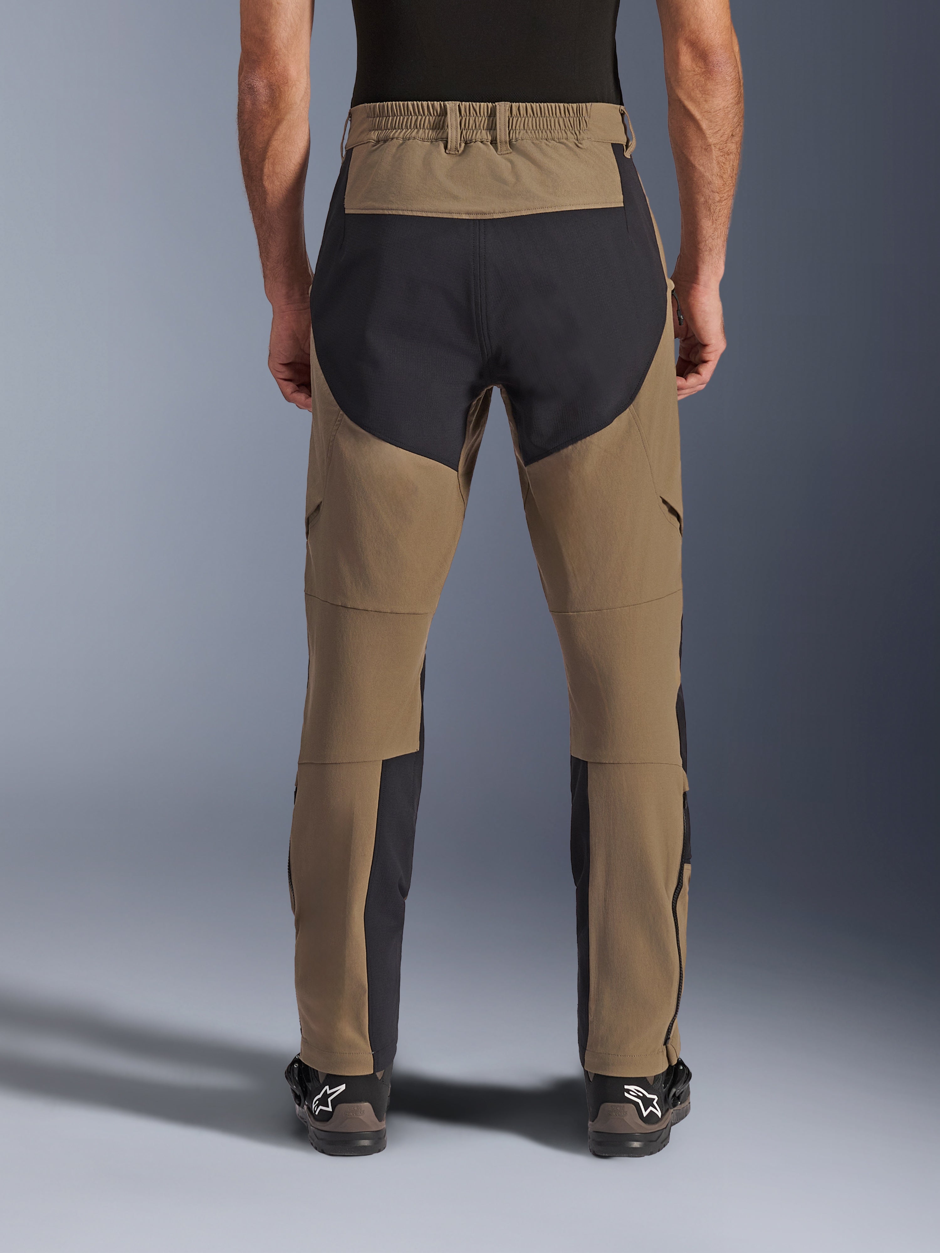 Alpinestars Flex-AST Explorer Pants, motocyklowe spodnie adventure, Walnut Black, widok z tyłu na motocykliście, czarne panele wzmacniające ripstop na siedzeniu i wewnętrznej stronie nogawek, elastyczny pas, długie zamki na dole nogawek, pasujące buty adventure