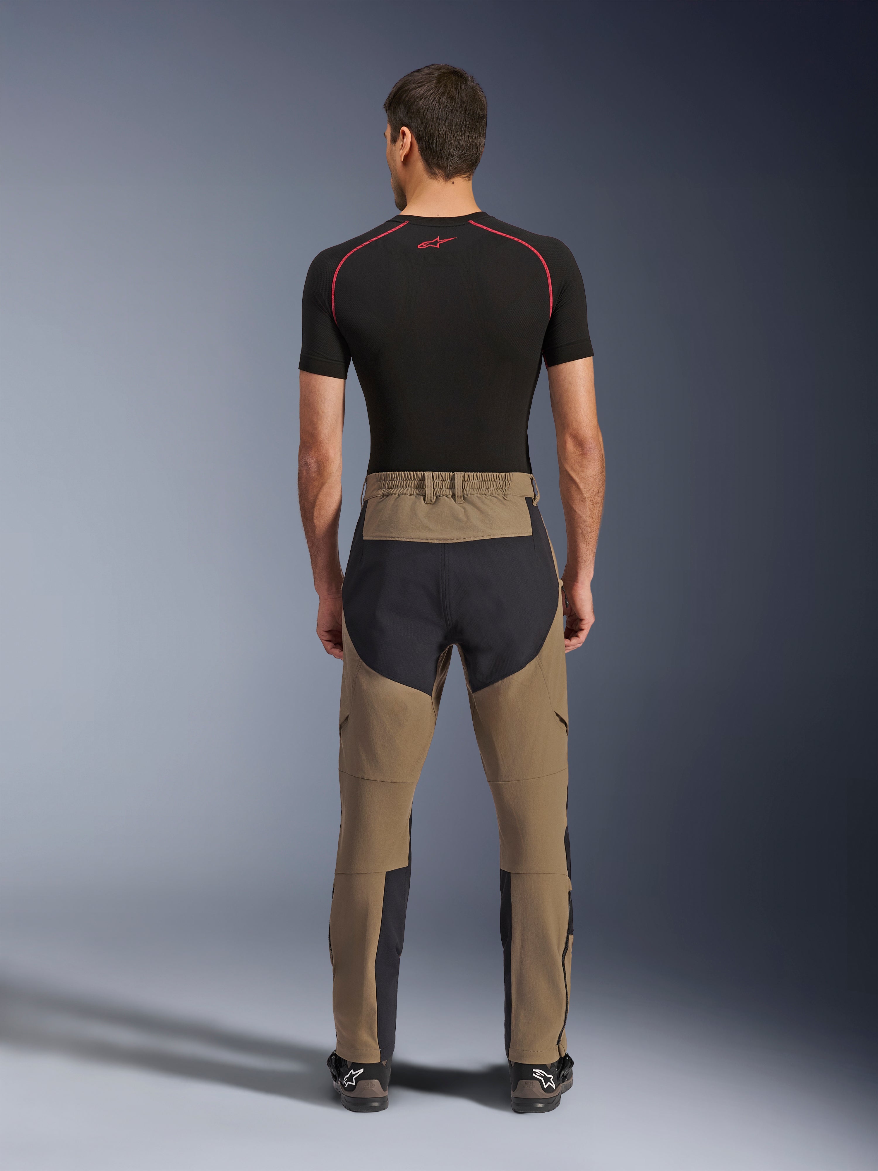 Spodnie Alpinestars Flex-AST Explorer, Walnut Black, widok z tyłu na modelu, wyposażone w wzmocniony czarny panel na siedzeniu i brązowy elastyczny materiał, zestawione z czarną warstwą bazową i technicznymi butami motocyklowymi, wytrzymała odzież do jazdy adventure