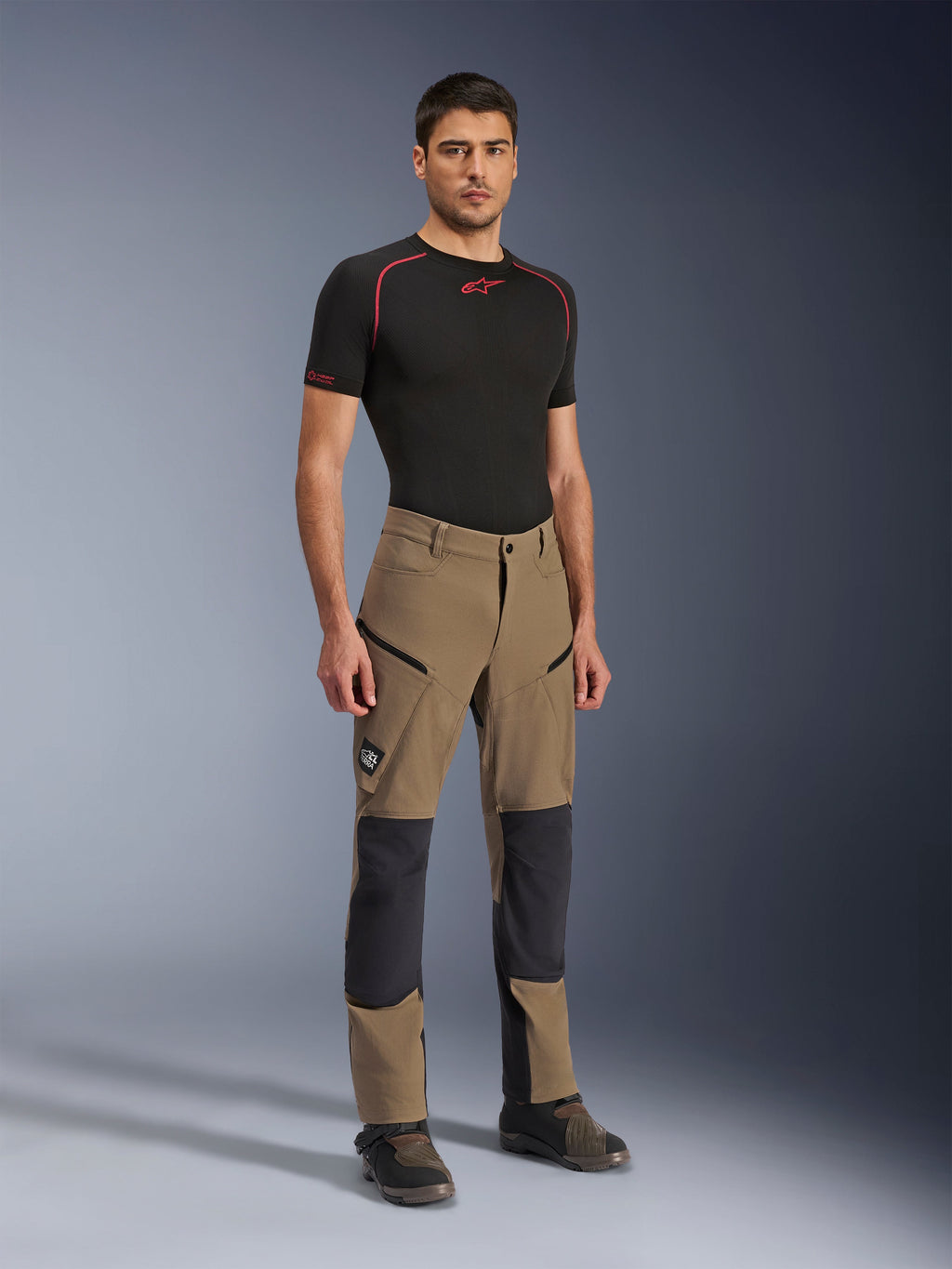Spodnie Alpinestars Flex-AST Explorer, motocyklowe spodnie adventure w kolorze Walnut Black, prezentowane przez modela w czarnej koszulce termoaktywnej z krótkim rękawem i butach adventure, wyposażone w kieszenie cargo na zamek i wzmocnione panele na kolanach, techniczna odzież dla motocyklistów dual-sport i turystycznych.