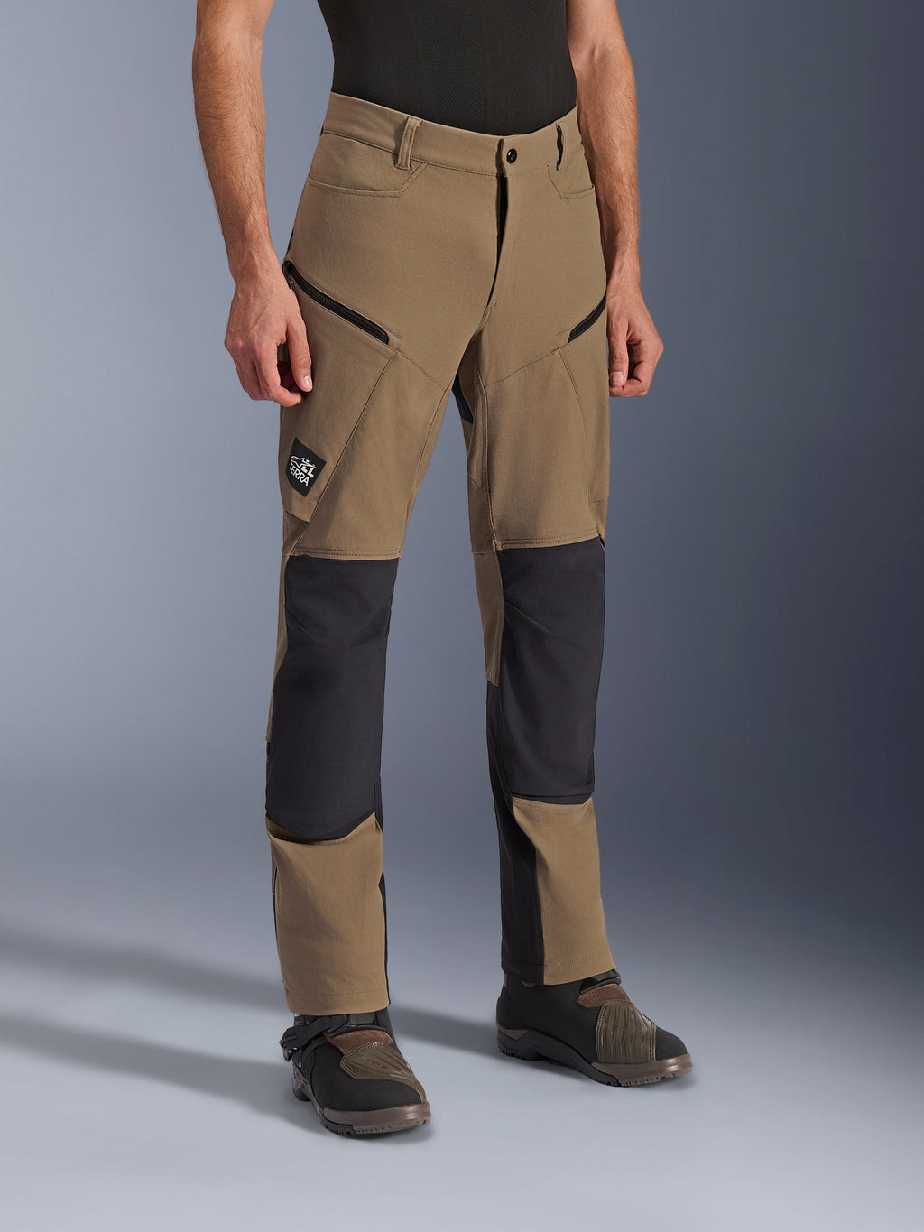 Spodnie Alpinestars Flex-AST Explorer, motocyklowe spodnie adventure w kolorze Walnut Black, beżowa tkanina z czarnymi wzmocnionymi panelami na kolanach i siedzeniu, kieszenie cargo na zamek, naszywka z logo All Terra, widoczne w zestawieniu z technicznymi butami motocyklowymi