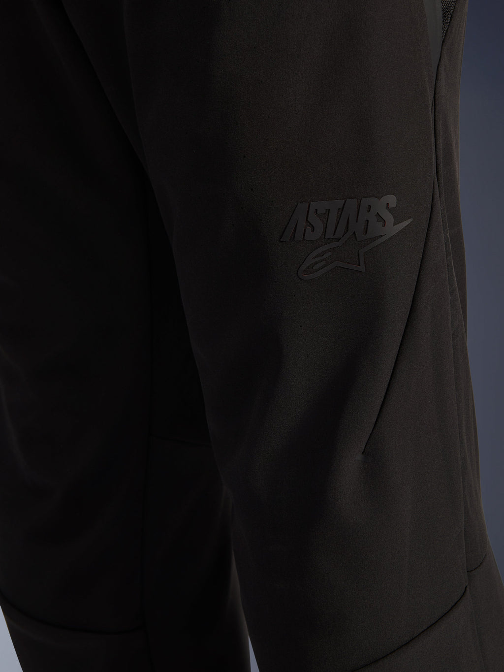 Spodnie Alpinestars Aeroshell Airflow, czarne spodnie motocyklowe, zbliżenie na obszar uda z wypukłym czarnym logo ASTARS, lekki, rozciągliwy materiał softshell z ergonomicznym układem szwów