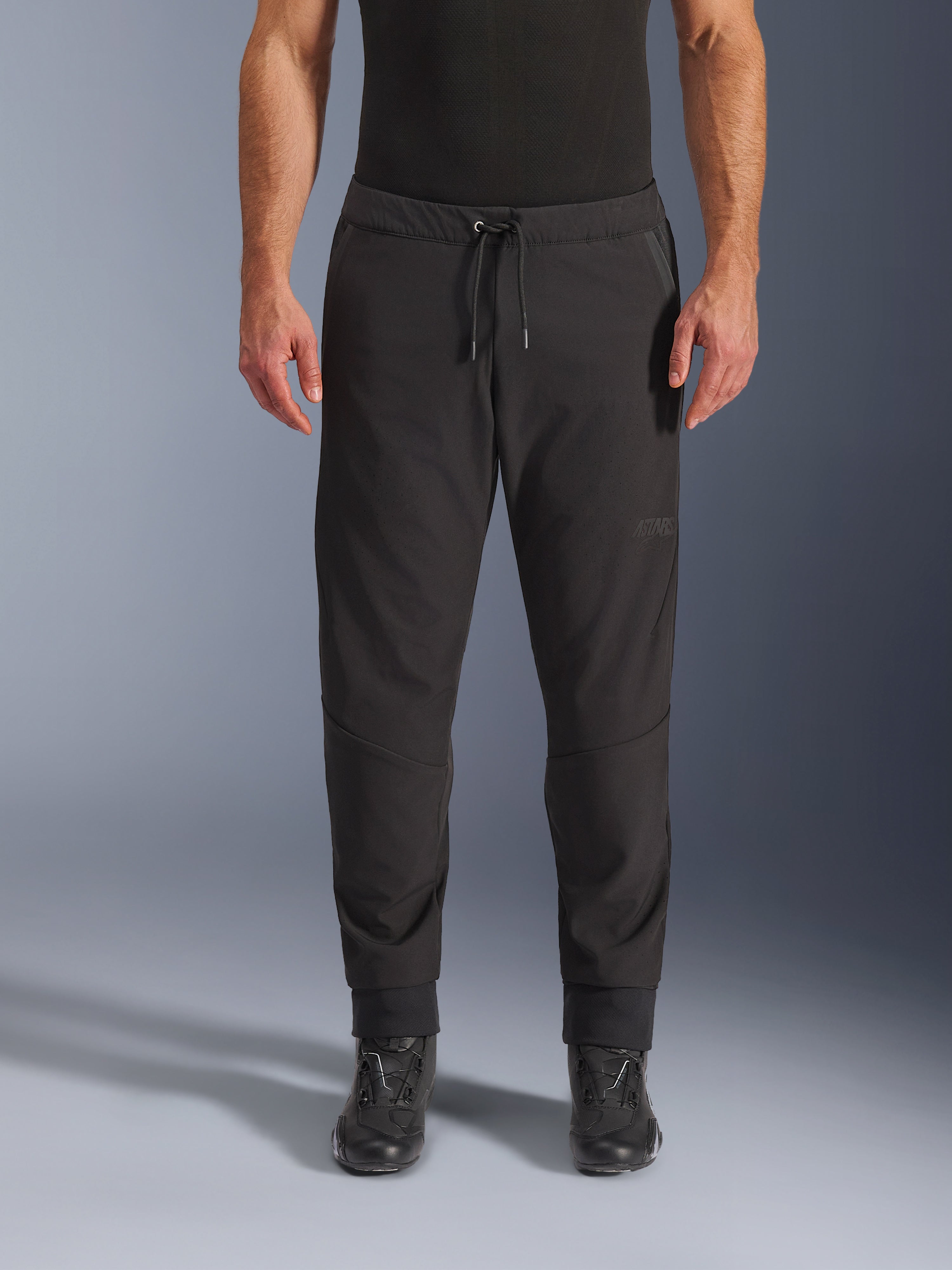 Spodnie Alpinestars Aeroshell Airflow, czarne techniczne spodnie motocyklowe, elastyczna konstrukcja softshell z laserowo perforowanymi strefami, pas ze sznurkiem i logo ASTARS, noszone z czarnymi butami, dla motocyklistów sportowych i turystycznych.