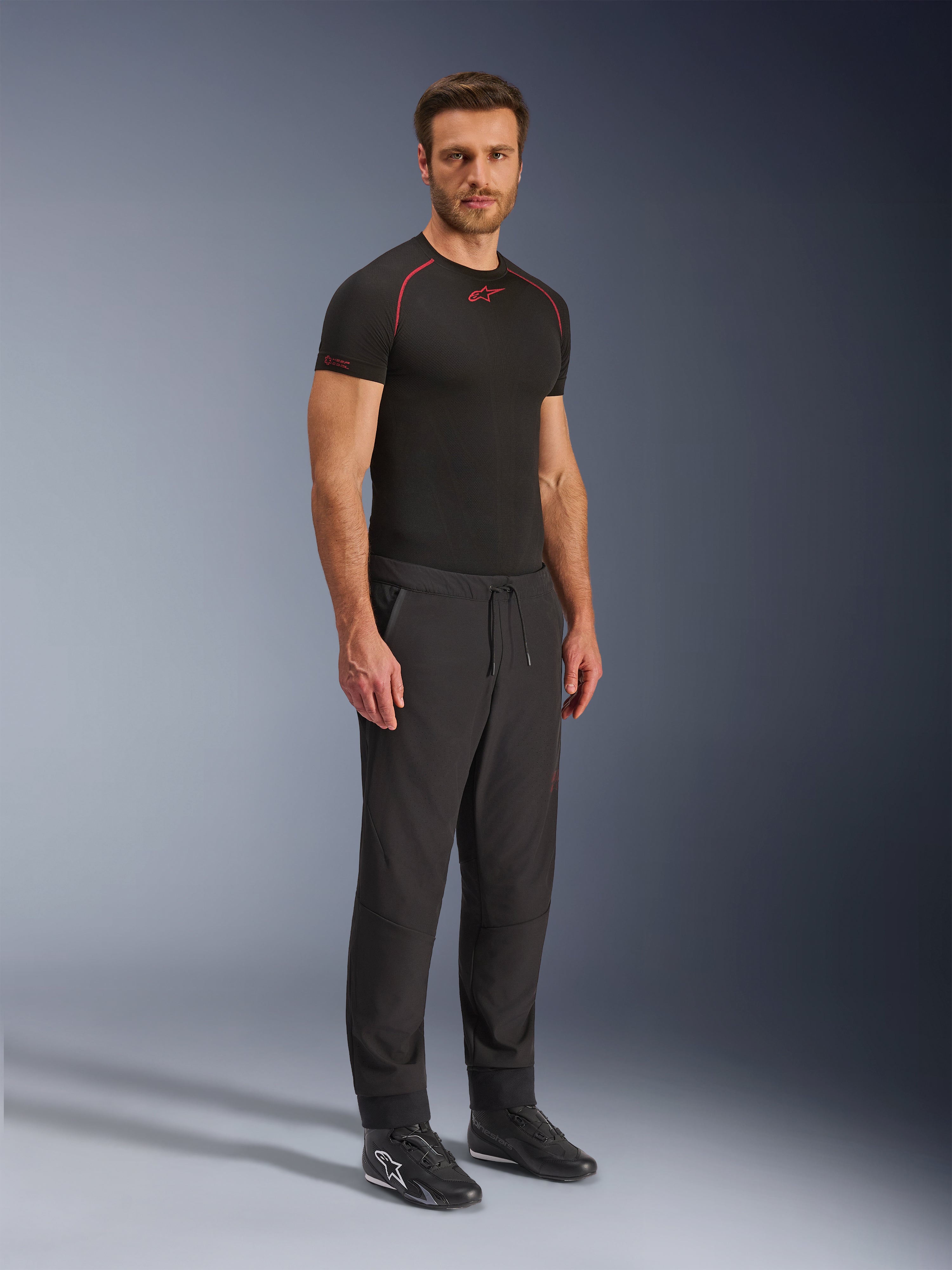 Alpinestars Aeroshell Airflow Pants, czarne spodnie motocyklowe, zdjęcie studyjne całej sylwetki modela ubranego w spodnie softshell z regulacją sznurkiem w pasie i elastycznymi mankietami, zestawione z czarną koszulką termoaktywną Alpinestars i czarnymi butami do jazdy.