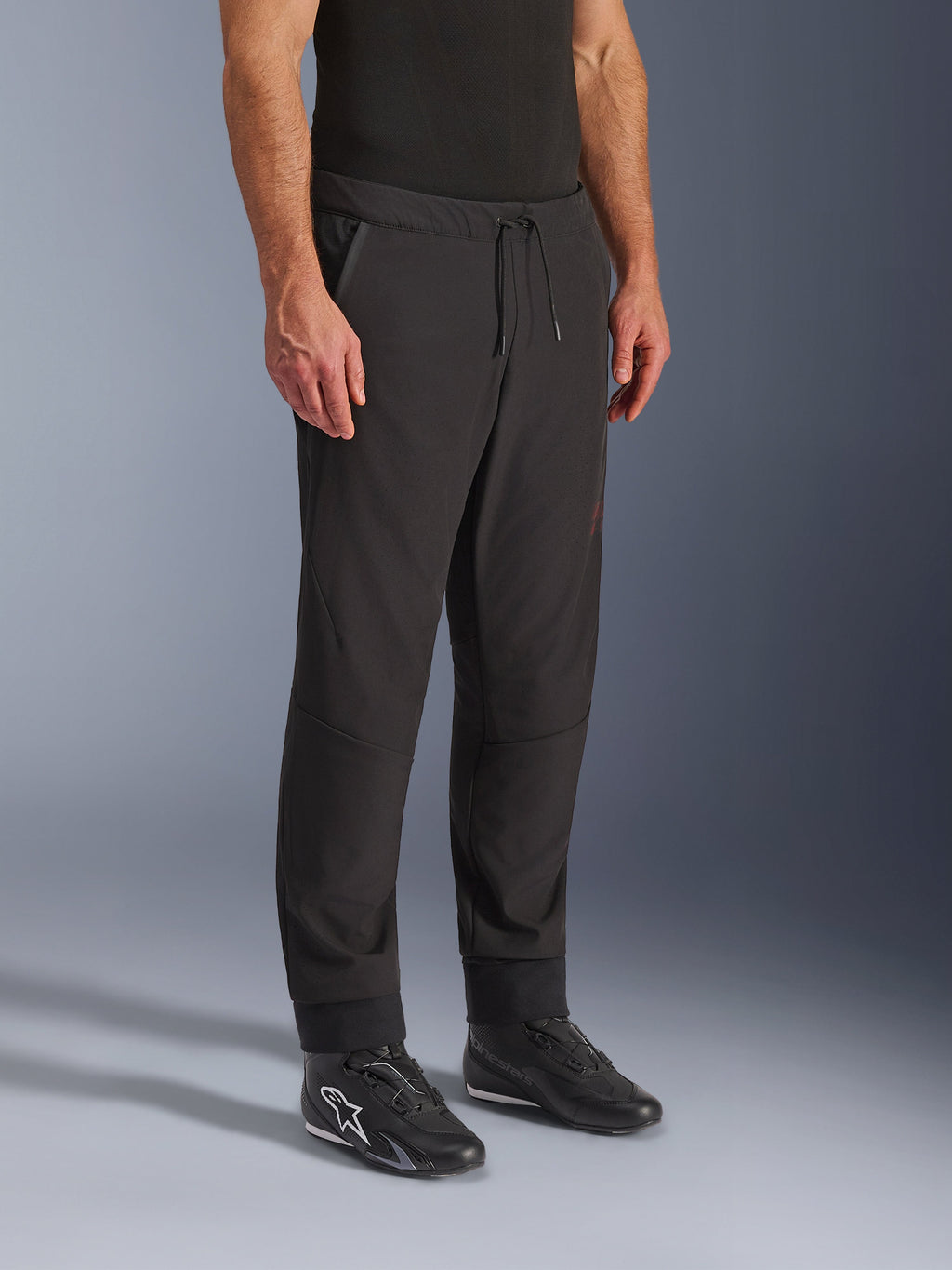 Alpinestars Aeroshell Airflow Pants, czarne spodnie motocyklowe, prezentowane przez modela z czarnymi butami motocyklowymi, wyposażone w sznurek w pasie, mankiety na kostkach i laserowo perforowany materiał softshell dla maksymalnej wentylacji