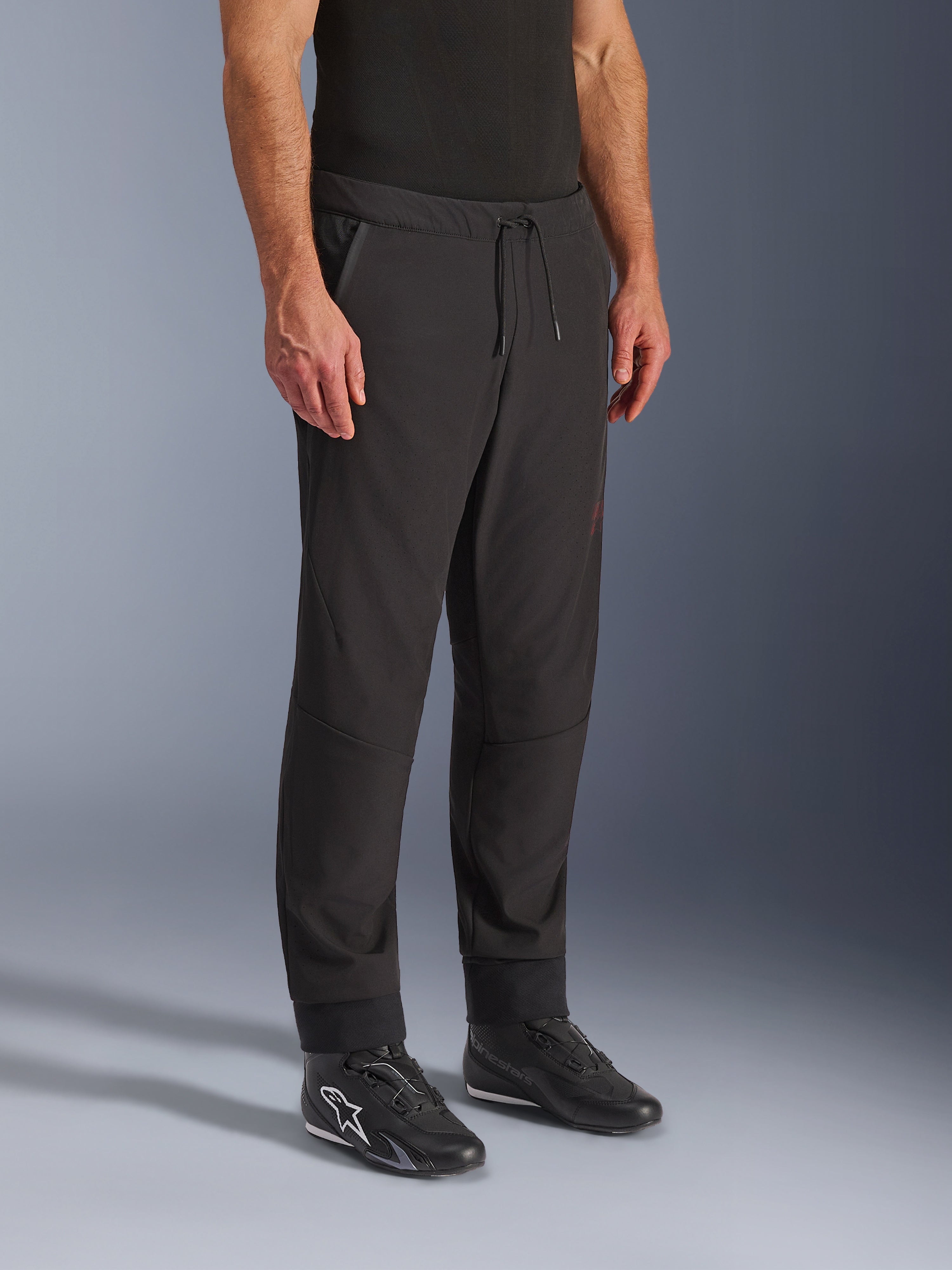 Alpinestars Aeroshell Airflow Pants, czarne spodnie motocyklowe, prezentowane przez modela z czarnymi butami motocyklowymi, wyposażone w sznurek w pasie, mankiety na kostkach i laserowo perforowany materiał softshell dla maksymalnej wentylacji