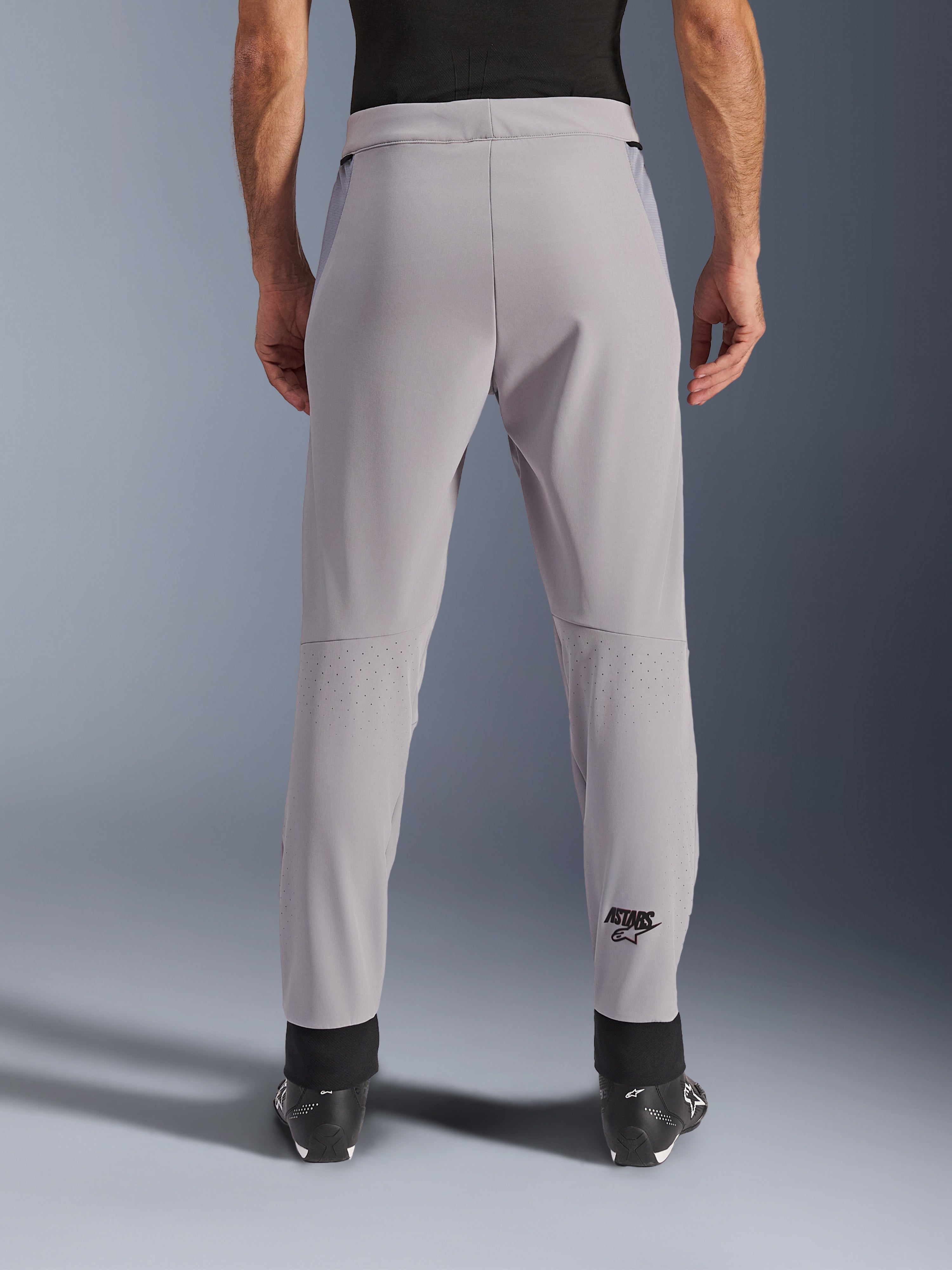 Alpinestars Aeroshell Airflow Pants, spodnie motocyklowe, Mid Gray, widok od tyłu ukazujący laserowo perforowane strefy na łydkach i czarne logo Astars, tekstylna konstrukcja softshell z czarnymi mankietami na kostkach i siatkowymi panelami na biodrach.