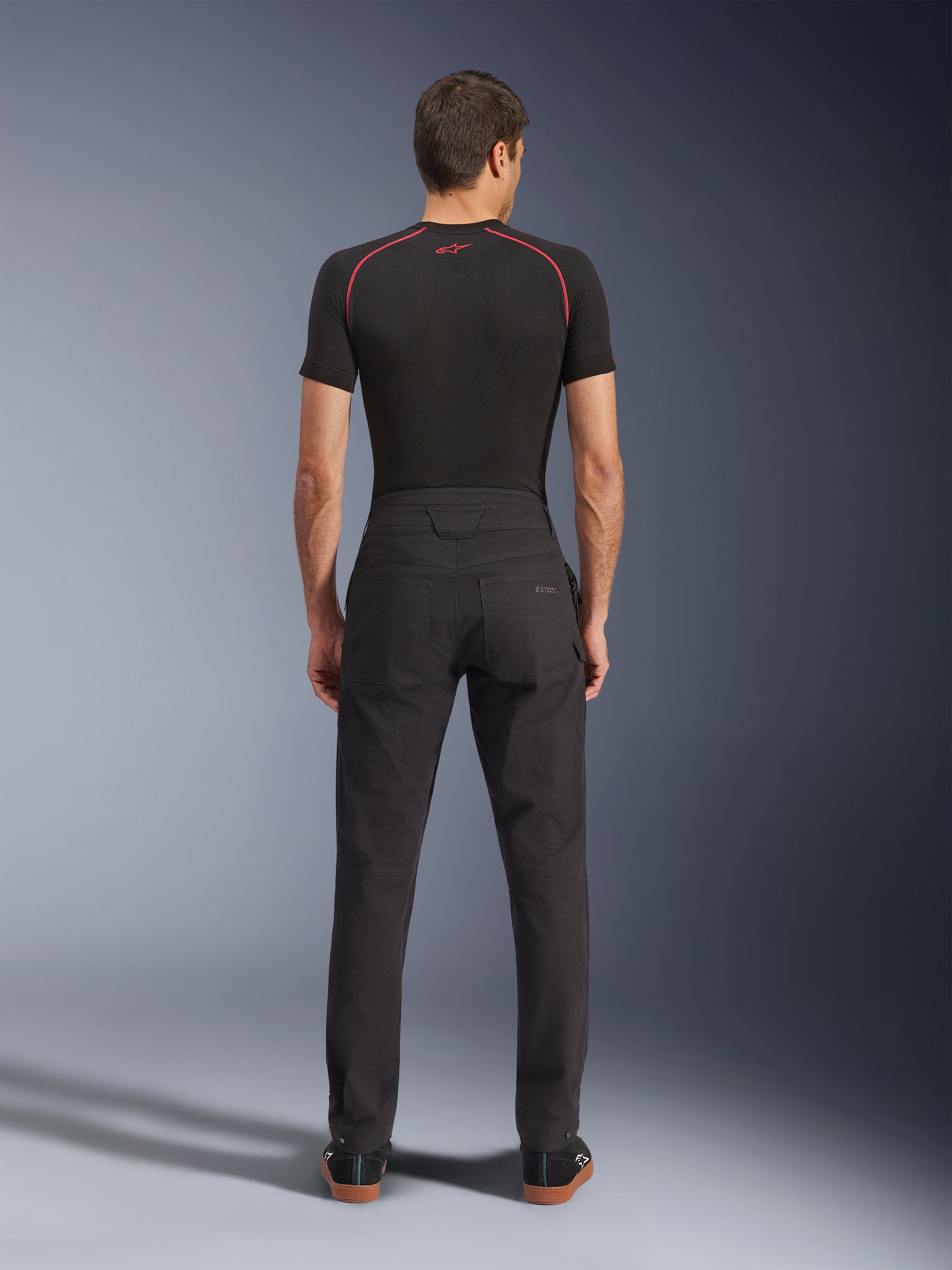 Alpinestars Flex-AST Canvas Pants, miejskie spodnie motocyklowe, czarne, widok z tyłu pokazujący design inspirowany odzieżą roboczą z tylnymi kieszeniami i podwyższonym tyłem, noszone z czarną warstwą bazową i sneakersami, ergonomiczny krój motocyklowy