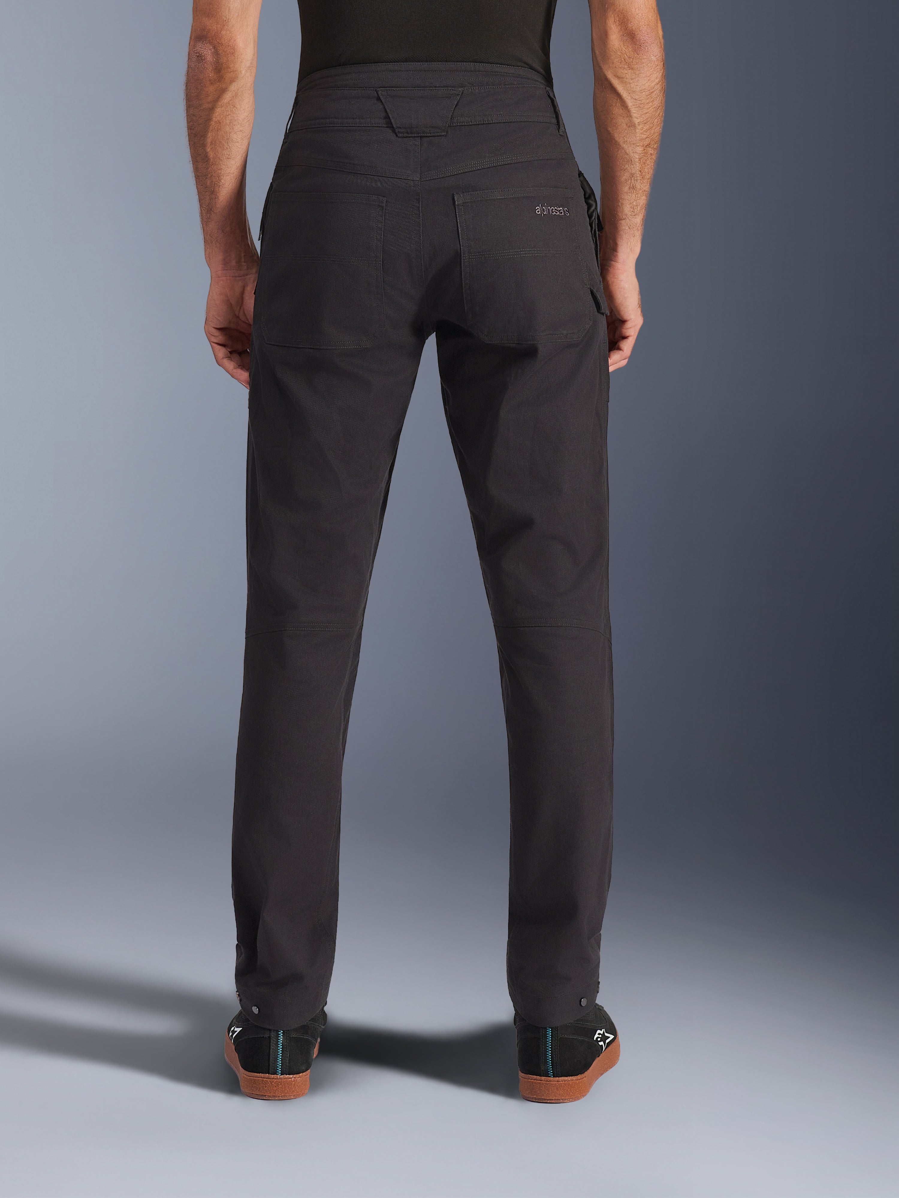 Spodnie Alpinestars Flex-AST Canvas, miejskie spodnie motocyklowe, czarne, widok z tyłu przedstawiający tylne kieszenie z haftowanym logo, podwyższony tył i ergonomiczne szwy na nogawkach, noszone z czarnym obuwiem codziennym