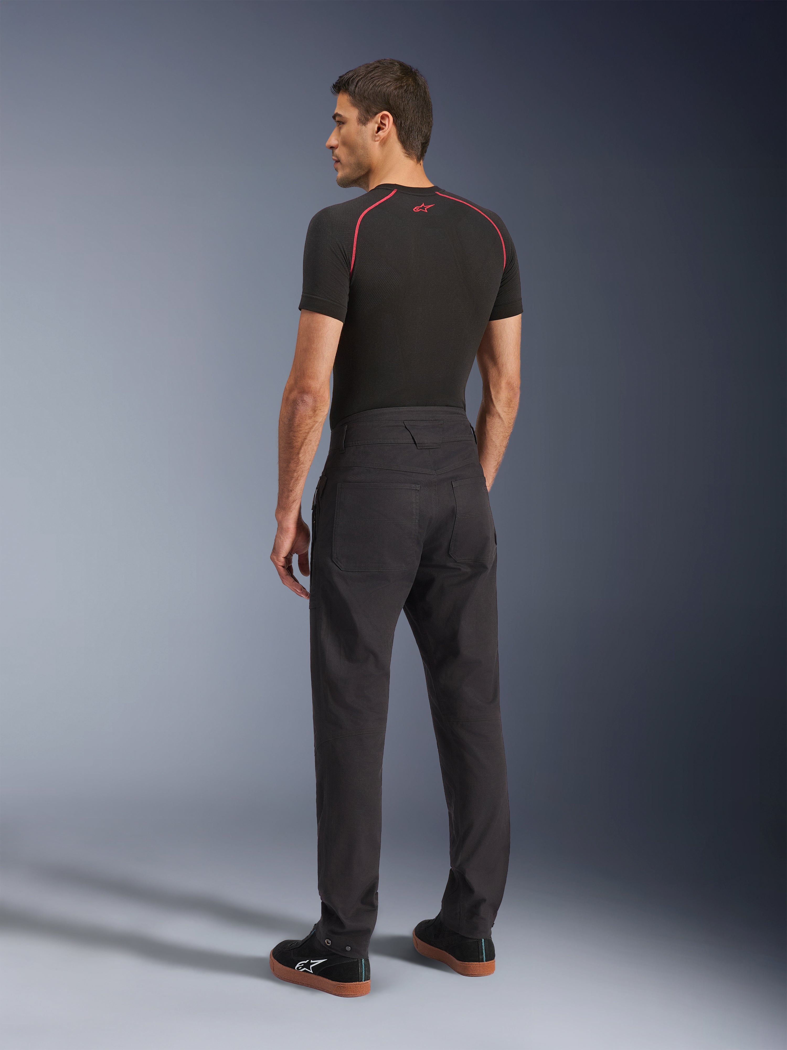 Spodnie Alpinestars Flex-AST Canvas, czarne miejskie spodnie motocyklowe, widok z tyłu na modelu, w zestawieniu z czarną warstwą bazową z krótkim rękawem i czarnymi sneakersami, wyróżniające się designem inspirowanym odzieżą roboczą z dużymi tylnymi kieszeniami i nowoczesnym kroju z prostymi nogawkami.