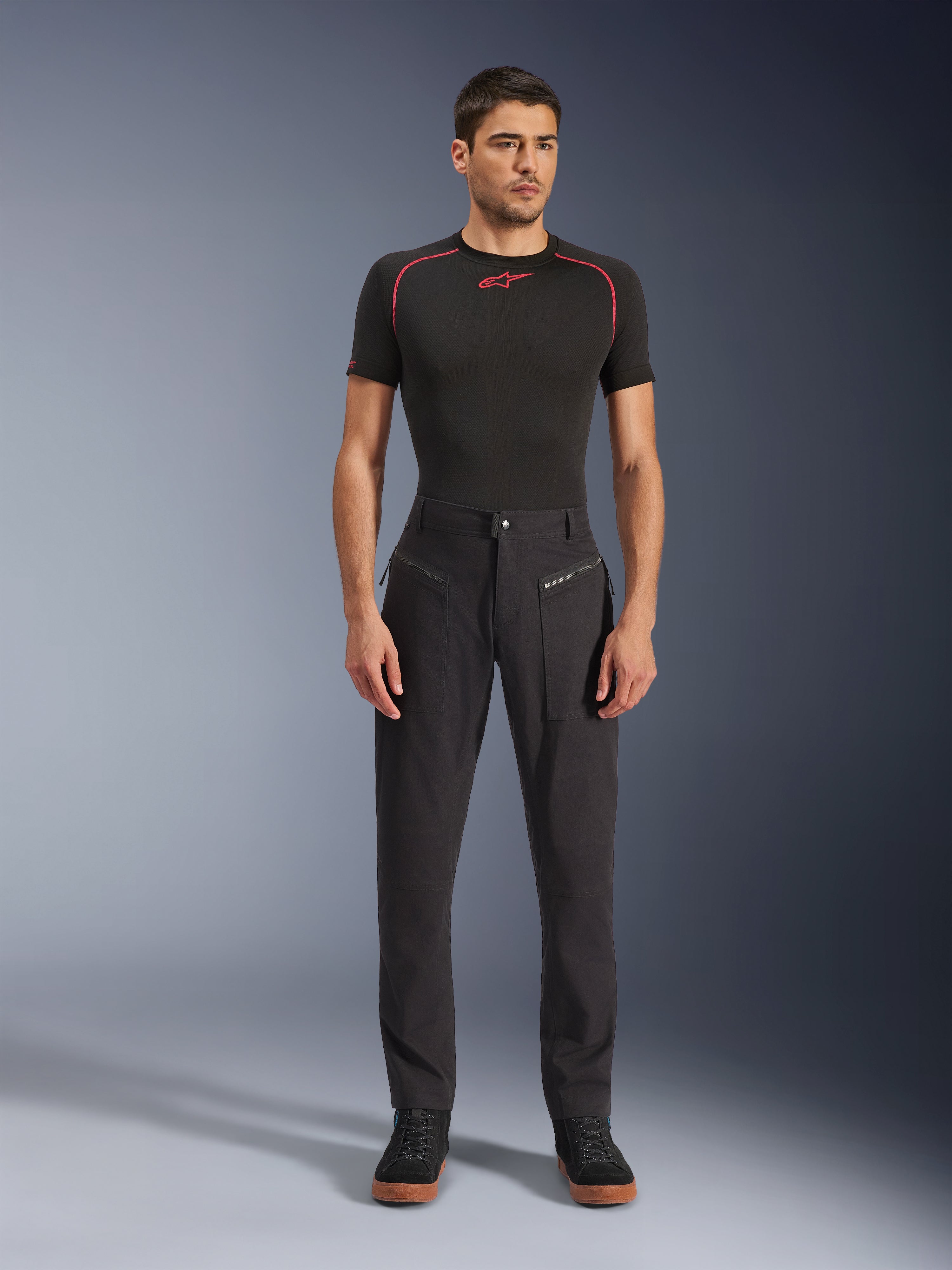 Spodnie Alpinestars Flex-AST Canvas, miejskie spodnie motocyklowe, czarne, prezentowane przez modela w czarnej warstwie bazowej z krótkim rękawem i czerwonymi przeszyciami oraz czarnych codziennych sneakersach, podkreślone przednie kieszenie na zamek i ergonomiczny design z materiału canvas.