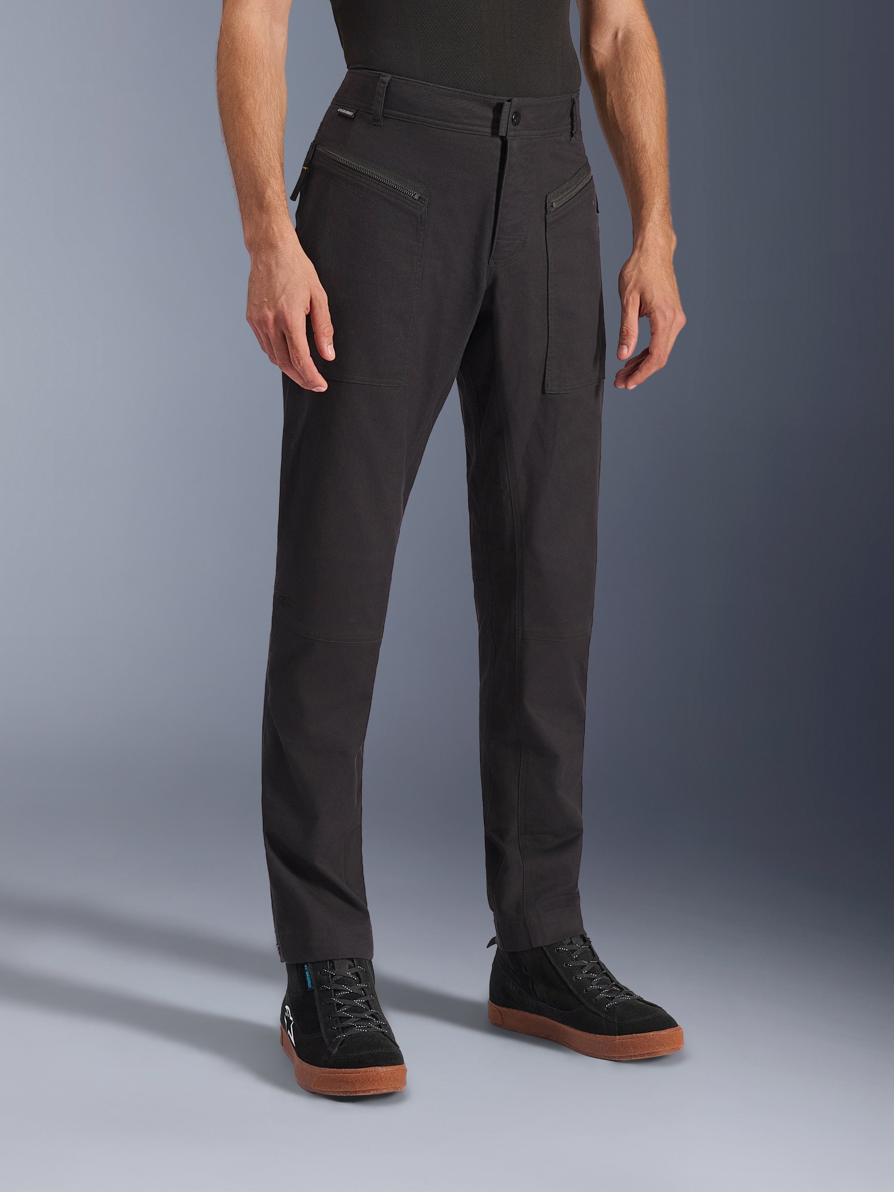 Spodnie Alpinestars Flex-AST Canvas, czarne, miejskie spodnie motocyklowe, prezentowane na modelu w czarnych sneakersach, tradycyjny design odzieży roboczej, elastyczna tkanina polibawełniana, funkcjonalne przednie kieszenie zapinane na zamek.