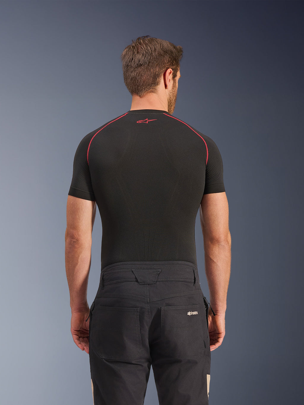 Alpinestars Flex-AST Canvas Pants w kolorze Black Aluminum, widok z tyłu na modelu, miejskie spodnie motocyklowe inspirowane odzieżą roboczą, logo na tylnej kieszeni, zestawione z czarną warstwą bazową z krótkim rękawem i czerwonymi przeszyciami.
