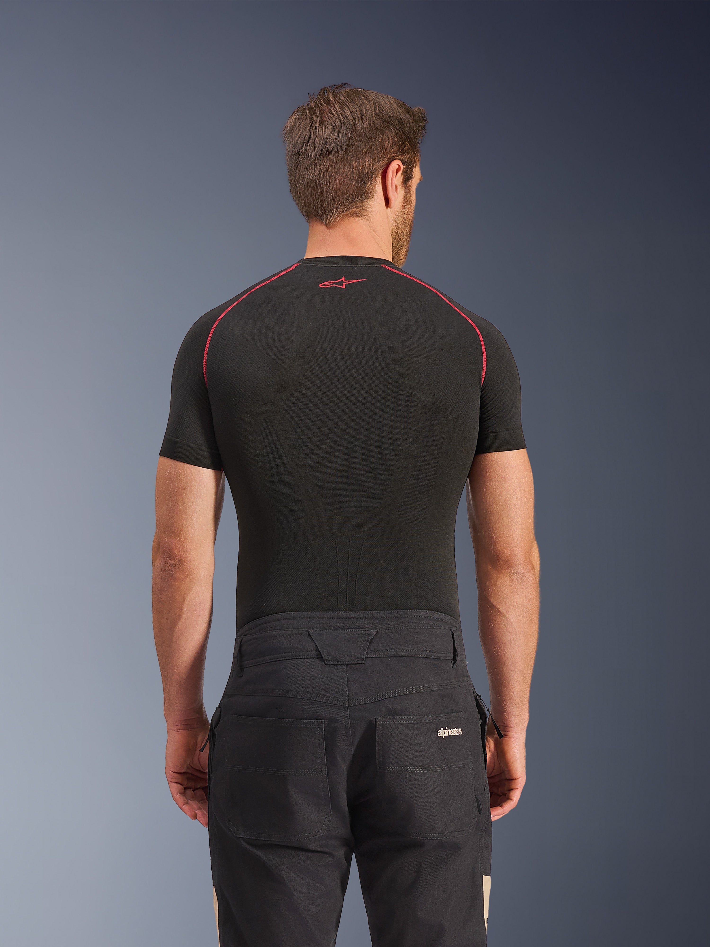 Alpinestars Flex-AST Canvas Pants w kolorze Black Aluminum, widok z tyłu na modelu, miejskie spodnie motocyklowe inspirowane odzieżą roboczą, logo na tylnej kieszeni, zestawione z czarną warstwą bazową z krótkim rękawem i czerwonymi przeszyciami.
