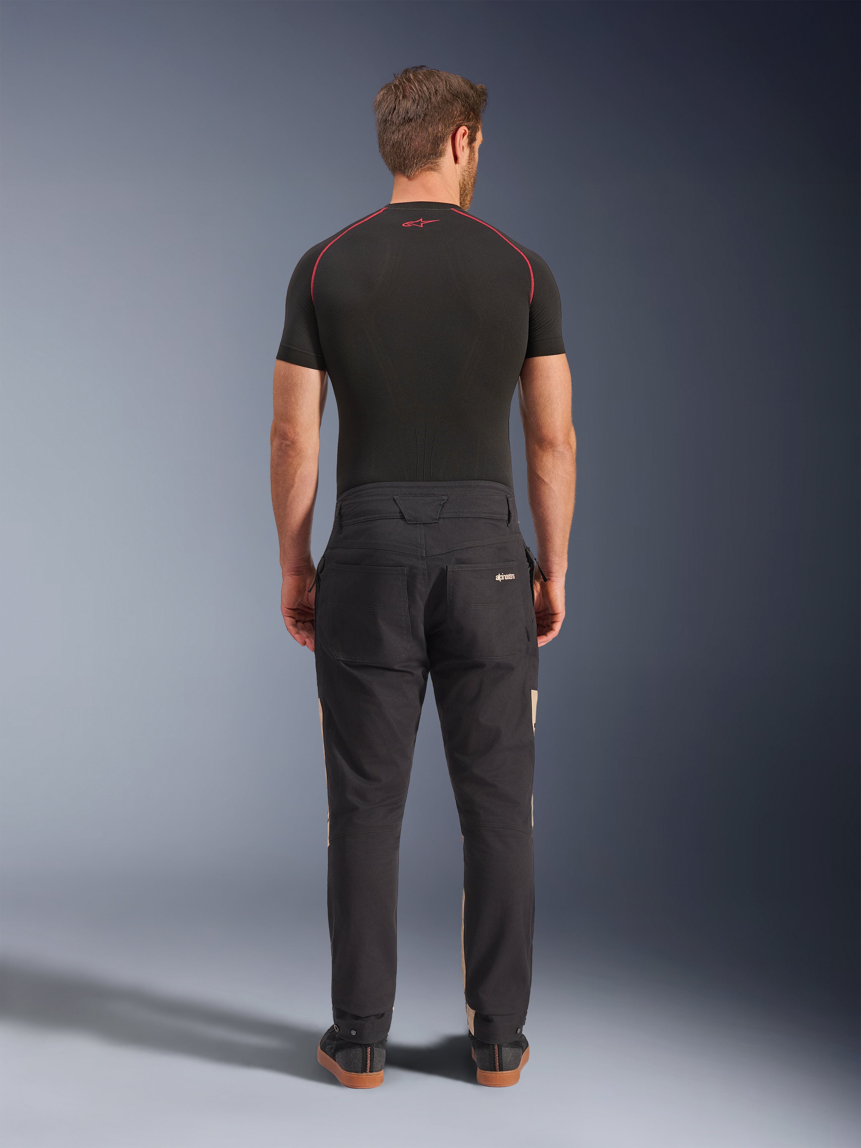 Alpinestars Flex-AST Canvas Pants w kolorze Black Aluminum, widok z tyłu na modelu, styl inspirowany odzieżą roboczą z tylnymi kieszeniami i subtelnymi beżowymi panelami bocznymi, zestawione z czarną warstwą termoaktywną i sneakersami do jazdy.