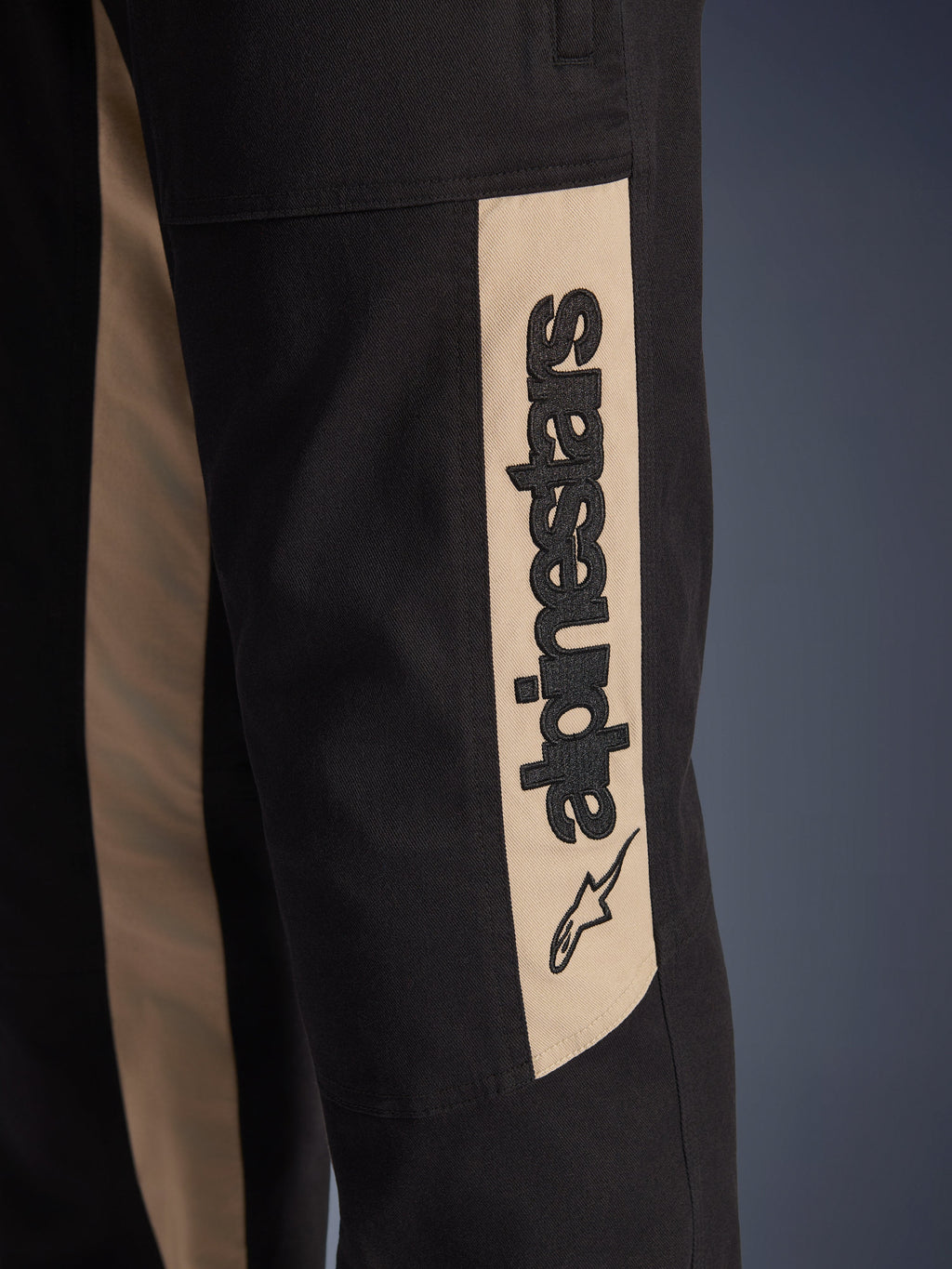Spodnie Alpinestars Flex-AST Canvas, zbliżenie na detal boku nogawki, czarne z beżowym panelem akcentującym Aluminum, pionowe czarne logo Alpinestars i ikona gwiazdy, wytrzymały twill z polibawełny do miejskiej jazdy motocyklowej