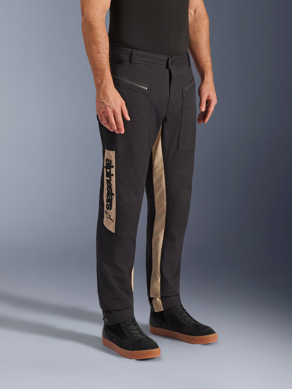 Alpinestars Flex-AST Canvas Pants, miejskie spodnie motocyklowe, kolor Black Aluminum, beżowe panele boczne z pionowym logo Alpinestars, design inspirowany odzieżą roboczą, kieszenie na zamek i ergonomiczny krój, noszone z czarnymi sneakersami dla uzyskania nowoczesnego, miejskiego wyglądu.