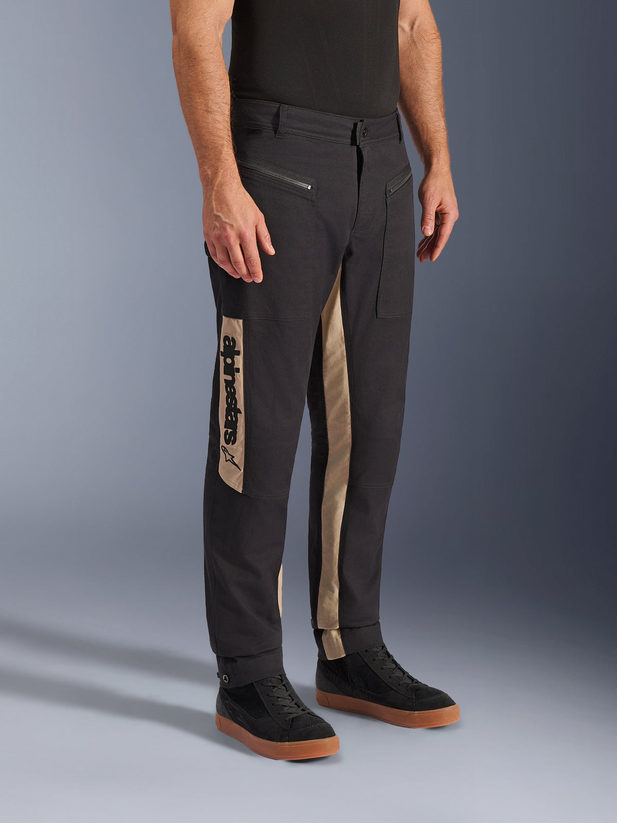 Alpinestars Flex-AST Canvas Pants, miejskie spodnie motocyklowe, kolor Black Aluminum, beżowe panele boczne z pionowym logo Alpinestars, design inspirowany odzieżą roboczą, kieszenie na zamek i ergonomiczny krój, noszone z czarnymi sneakersami dla uzyskania nowoczesnego, miejskiego wyglądu.