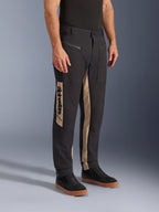 Alpinestars Flex-AST Canvas Pants, miejskie spodnie motocyklowe, kolor Black Aluminum, beżowe panele boczne z pionowym logo Alpinestars, design inspirowany odzieżą roboczą, kieszenie na zamek i ergonomiczny krój, noszone z czarnymi sneakersami dla uzyskania nowoczesnego, miejskiego wyglądu.