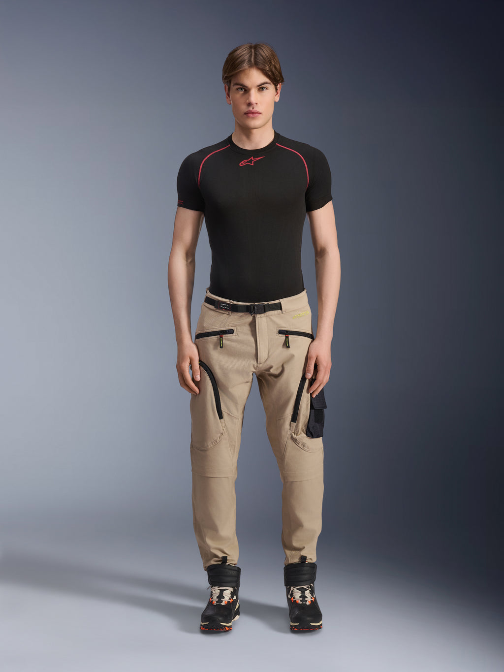 Męski model ubrany w spodnie Alpinestars Acteon w kolorze piaskowym (Sand), zestawione z czarną warstwą bazową z krótkim rękawem i czarnymi butami technicznymi, z kieszeniami cargo z czarnymi zamkami i zwężanym krojem, nowoczesna miejska odzież motocyklowa