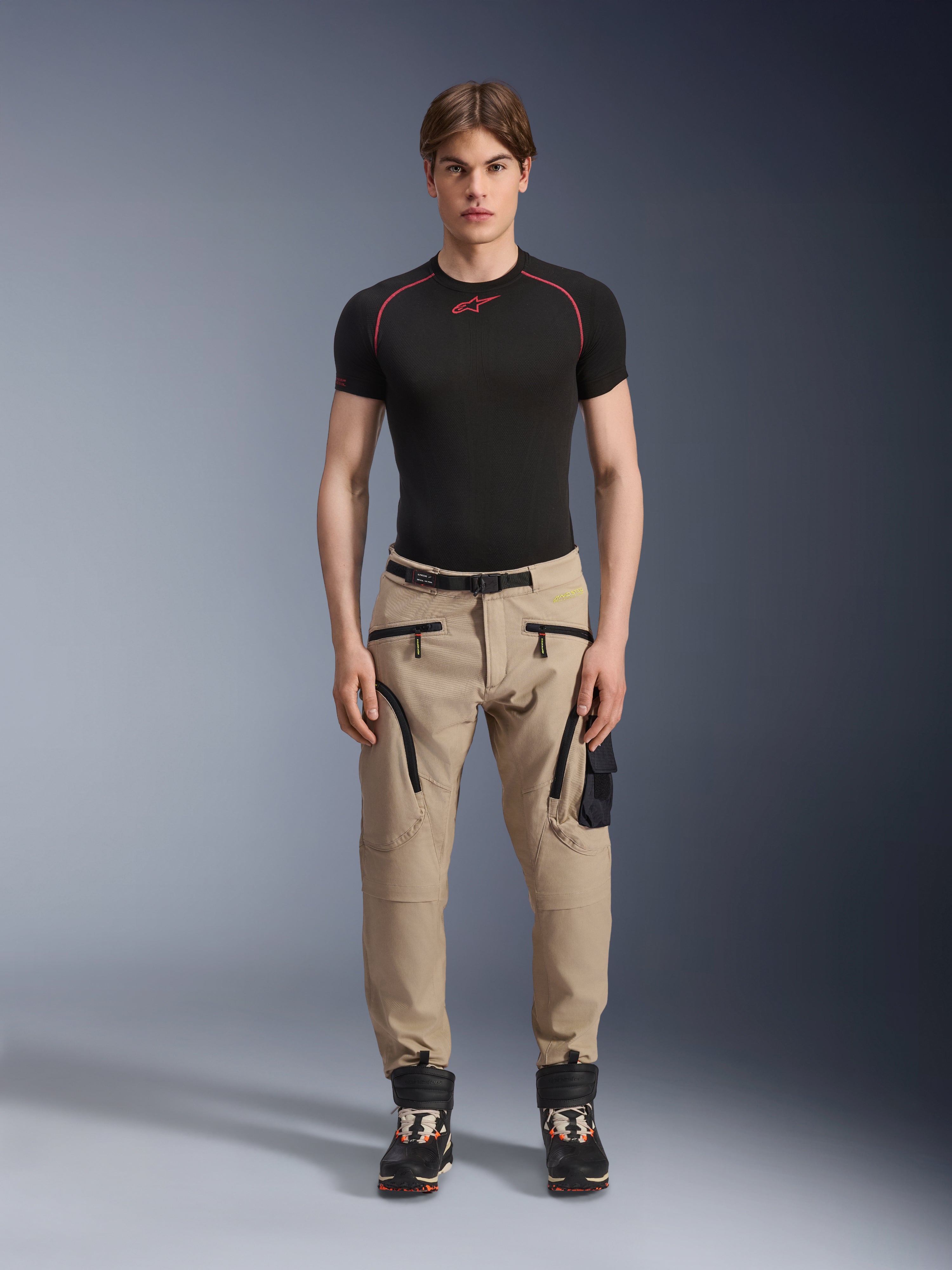 Męski model ubrany w spodnie Alpinestars Acteon w kolorze piaskowym (Sand), zestawione z czarną warstwą bazową z krótkim rękawem i czarnymi butami technicznymi, z kieszeniami cargo z czarnymi zamkami i zwężanym krojem, nowoczesna miejska odzież motocyklowa