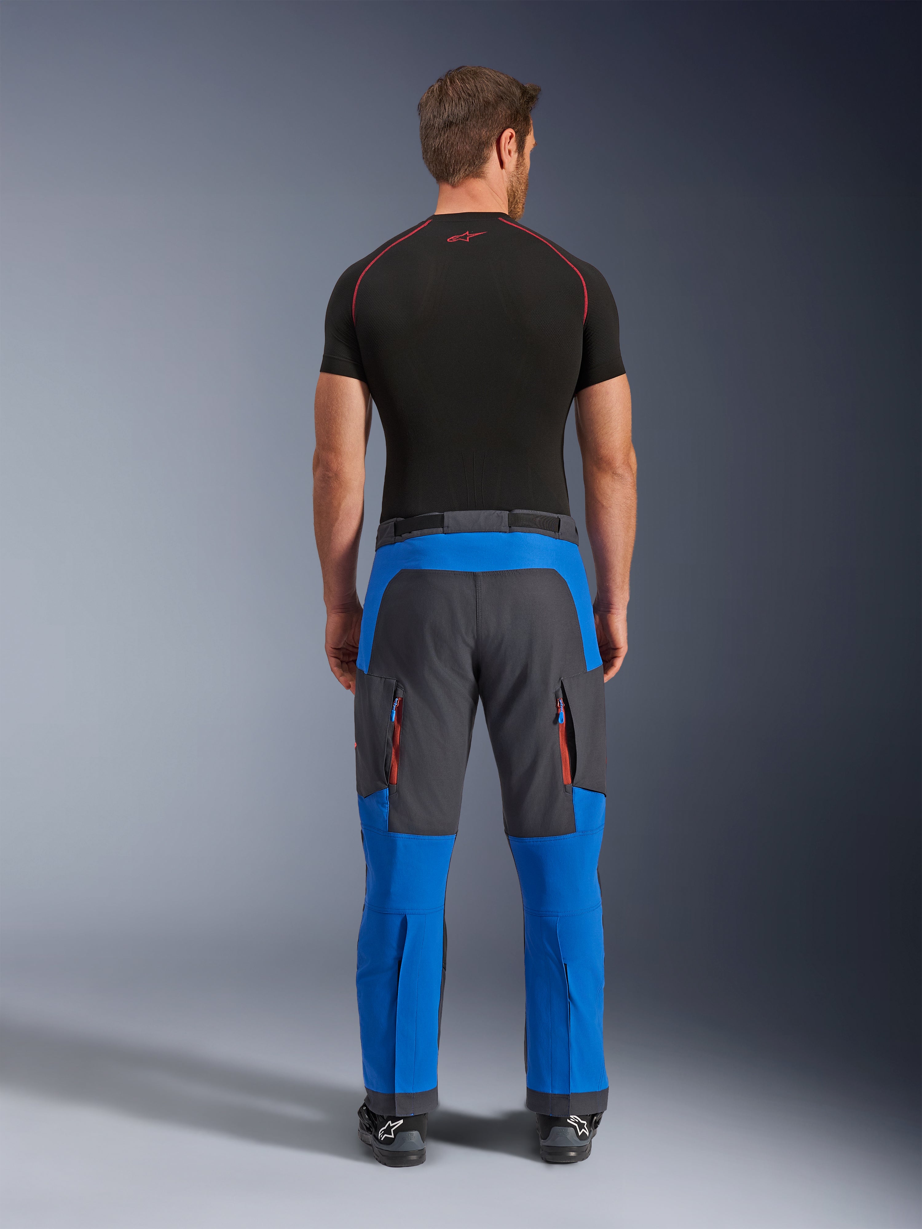 Alpinestars Maxdura Dual Pants w kolorze niebieskim i ciemnoszarym, motocyklowe spodnie adventure widoczne od tyłu, kieszenie cargo na zamek z pomarańczowymi detalami, krój na buty, zestawione z czarną warstwą bazową z krótkim rękawem.