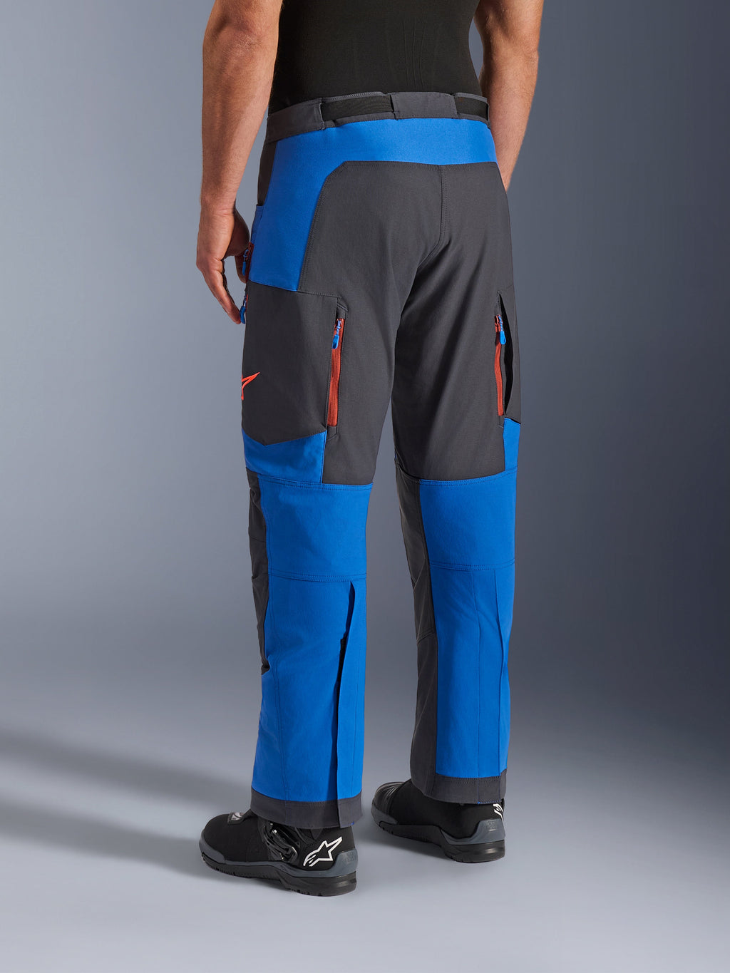 Alpinestars Maxdura Dual Pants, niebieskie i ciemnoszare, widok z tyłu przedstawiający kieszenie na zamek i strefy wentylacyjne, krój na buty noszony z czarnymi butami Alpinestars, wytrzymała odzież do jazdy adventure i dual-sport